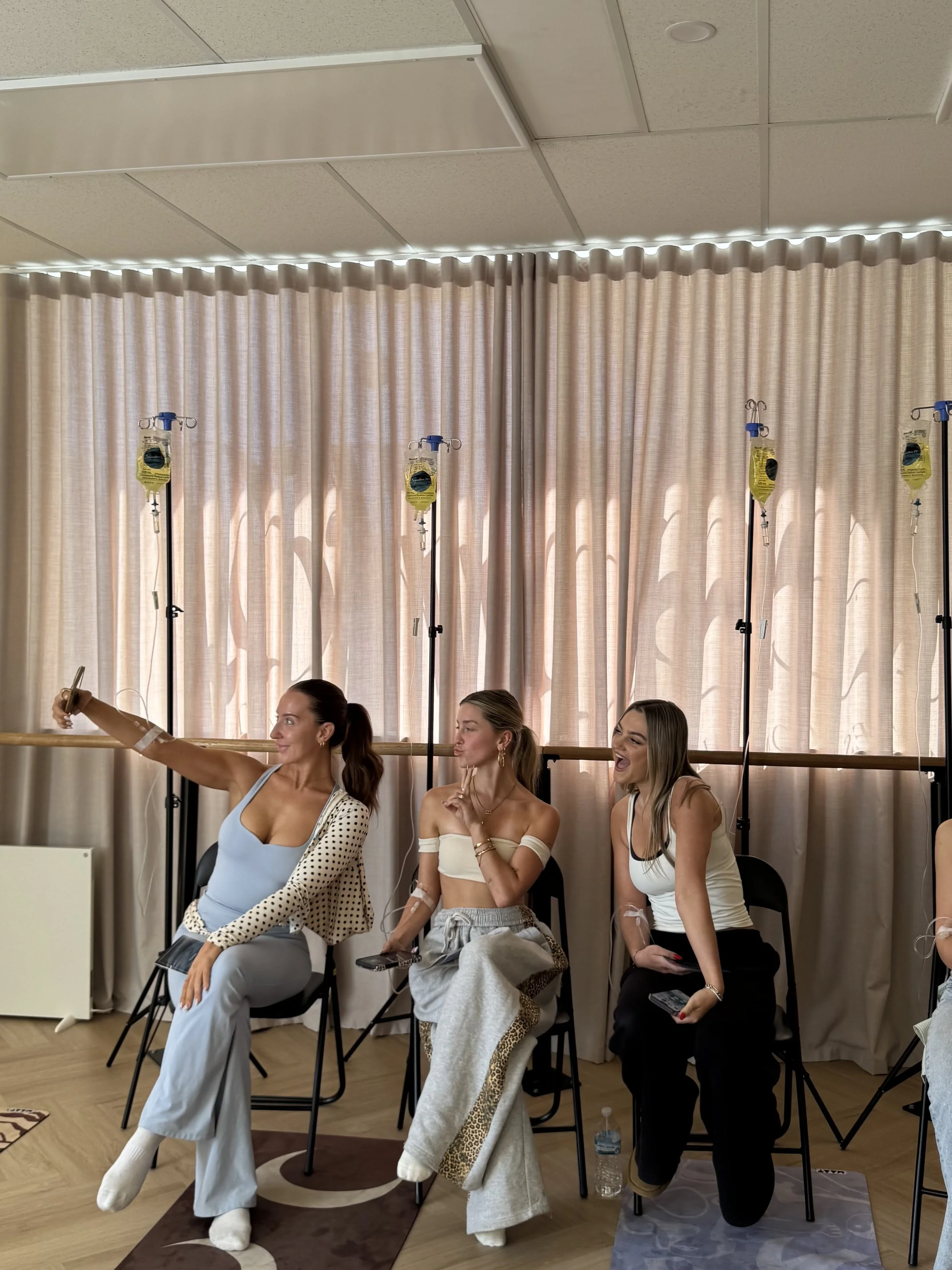 Three women participating in a wellness or meditation session, seated on chairs with IV drips attached, in a room with beige curtains and a wooden floor.