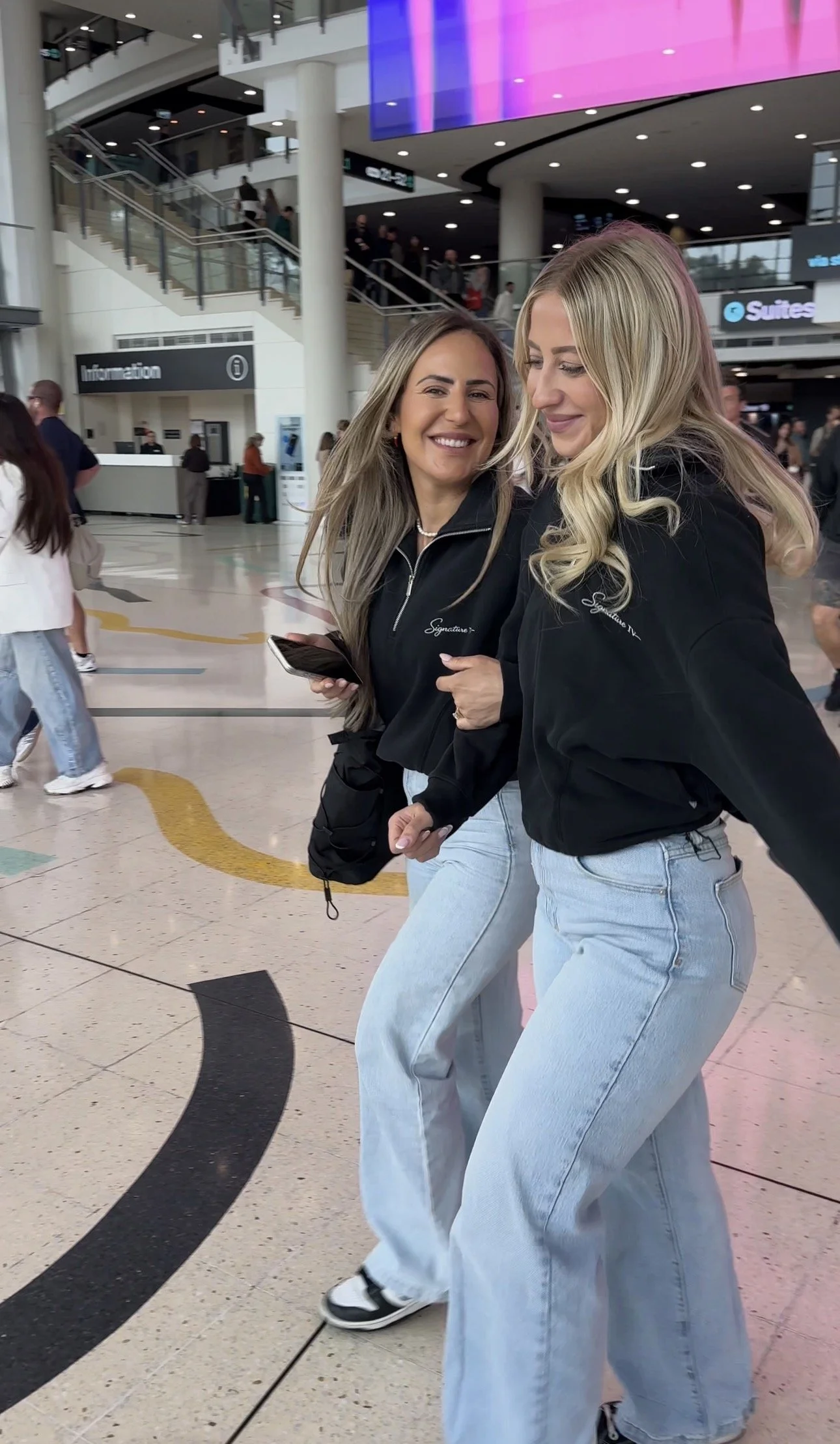 Two women with long hair, wearing black jackets and light blue jeans, walking inside an airport terminal. One woman is smiling and holding a phone, while the other is looking down with a gentle smile. The background shows multiple people, escalators, and signage.