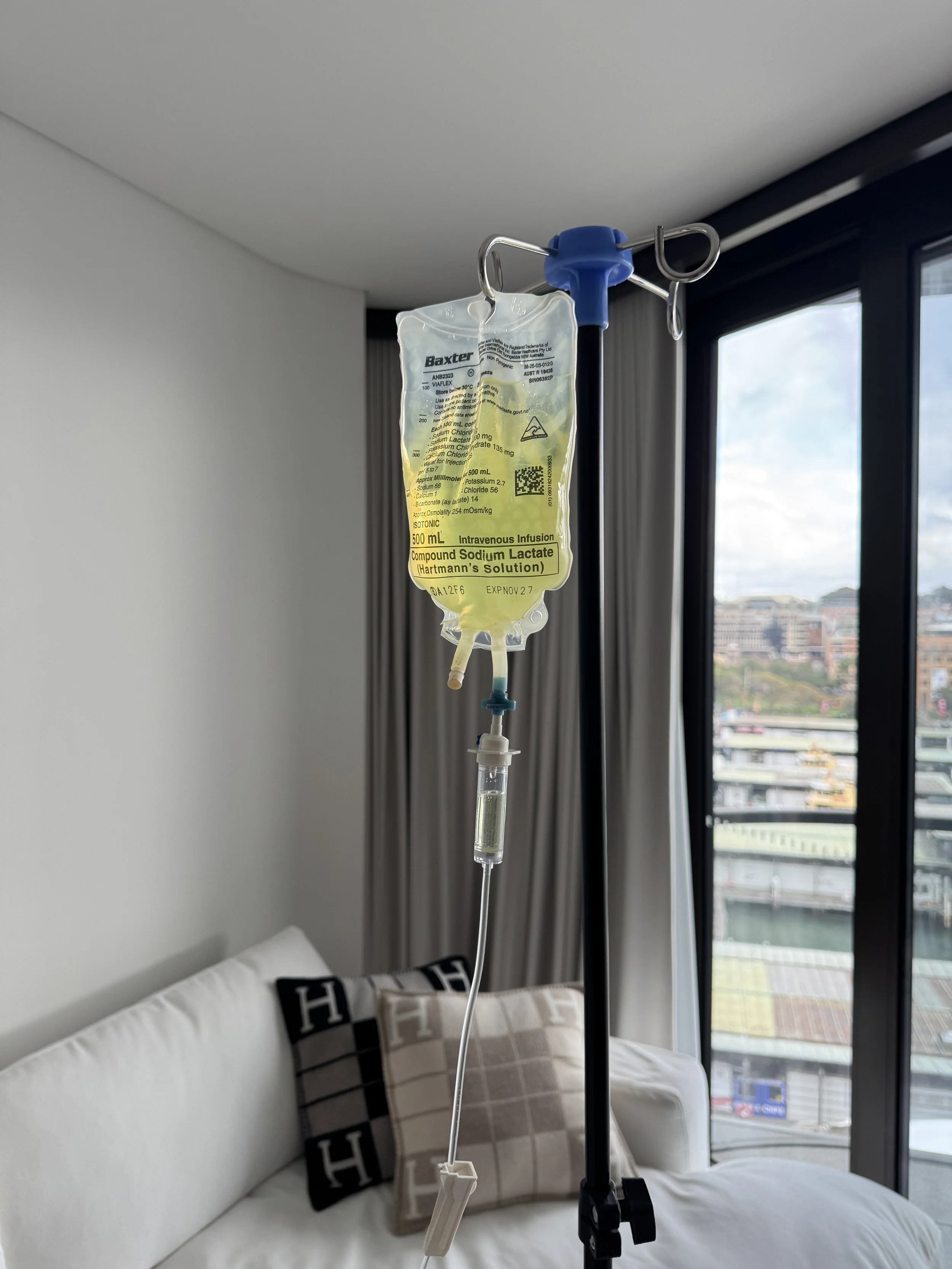 Hospital IV drip hanging in a bedroom with a bed, pillows, and large windows showing an urban cityscape.