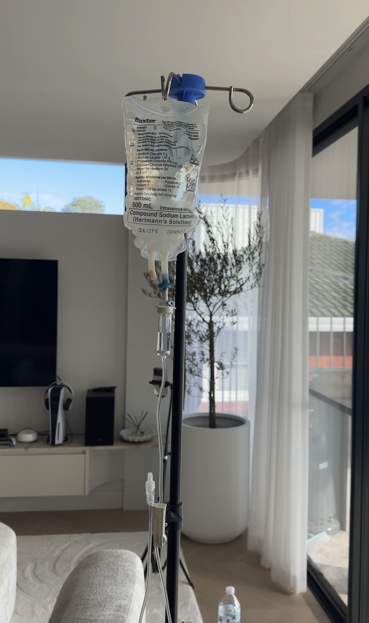 Intravenous (IV) drip bag hanging from a stand in a living room with a TV, plants, and curtains in the background.