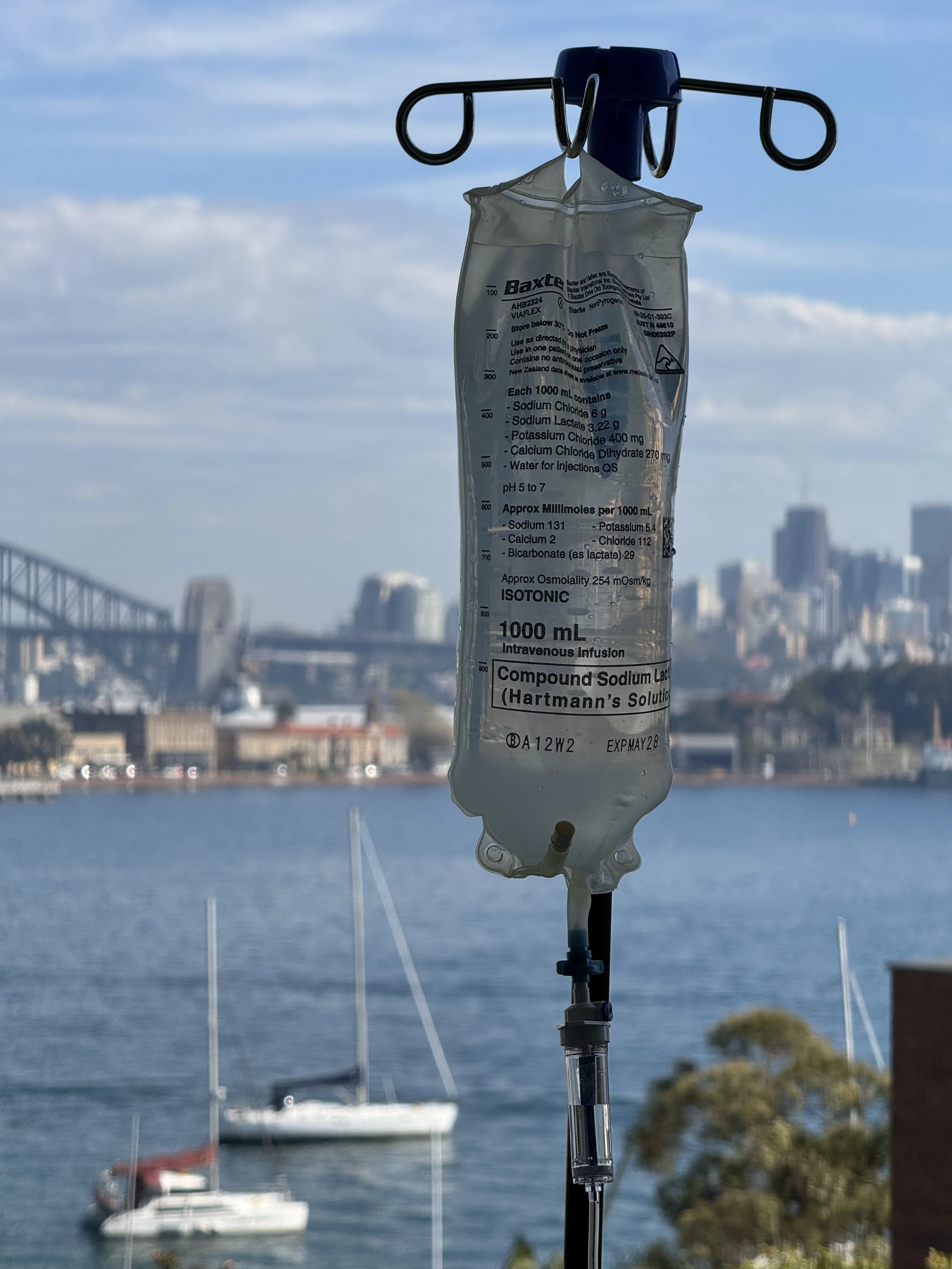 An intravenous drip bag floating in the air with a cityscape and water in the background.