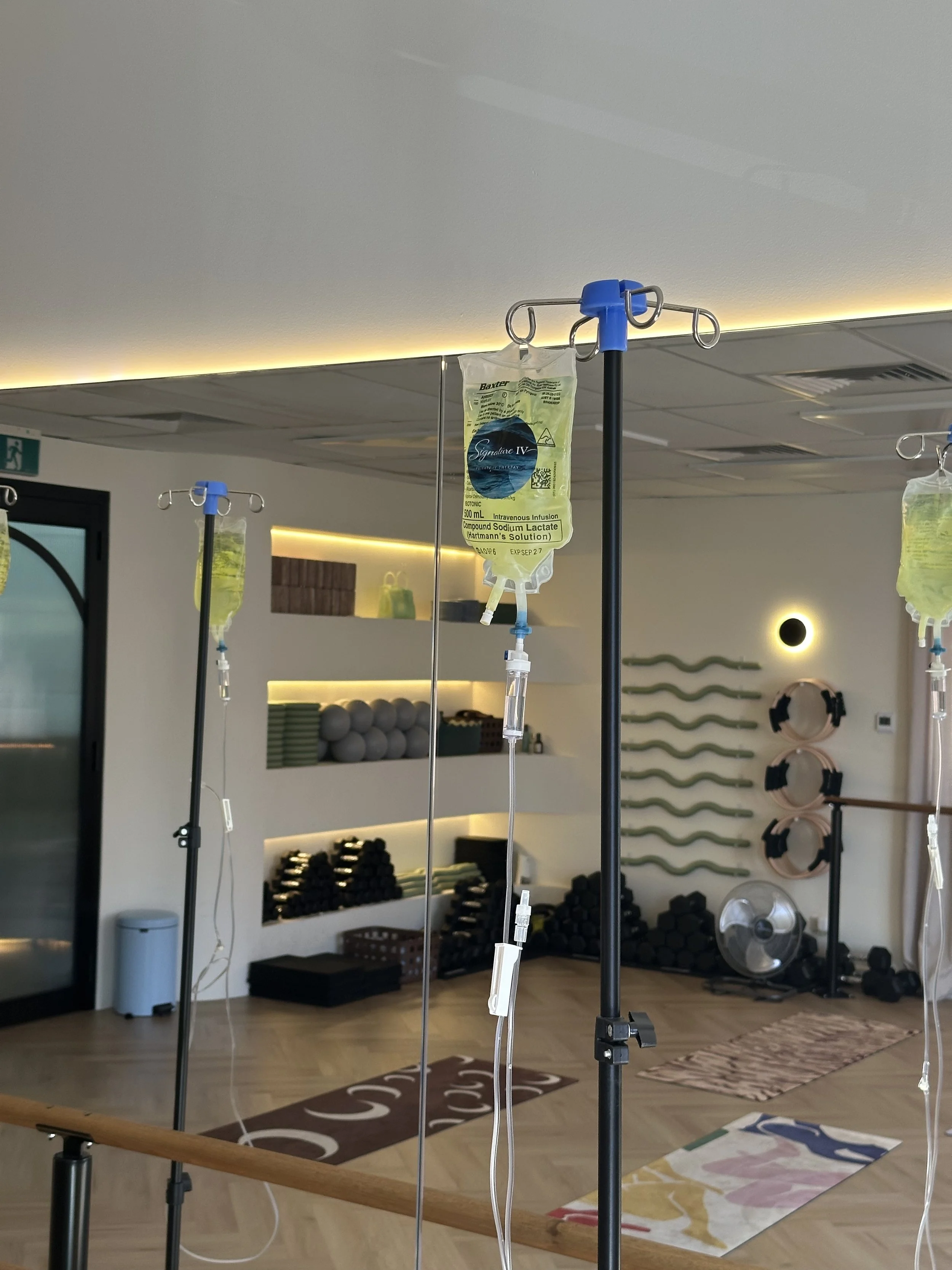 Medical IV drip hanging from a stand in a fitness or rehabilitation studio with yoga mats, exercise equipment, and a mirror in the background.