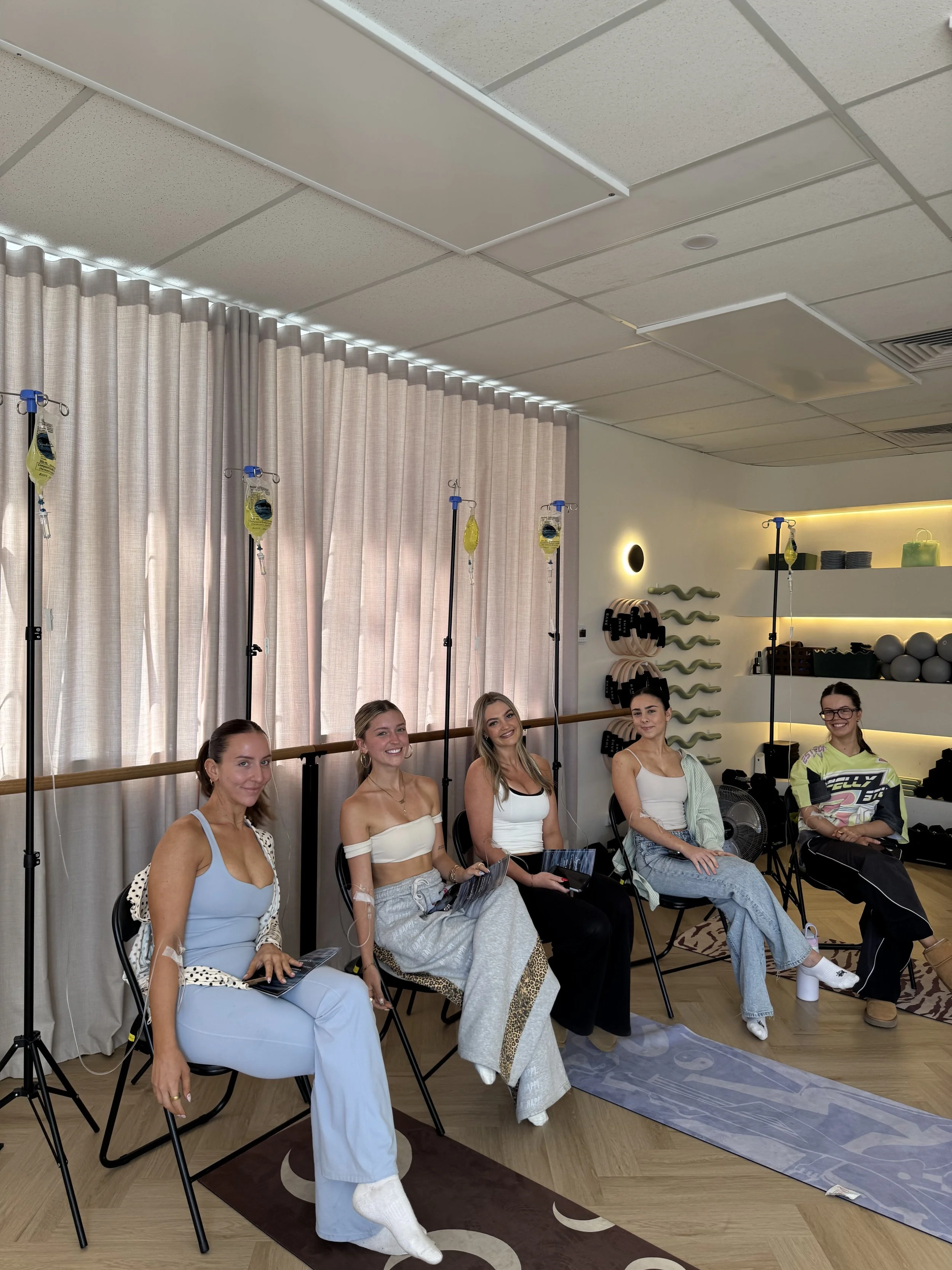 A group of five women sitting in chairs, smiling and holding papers, in a room with exercise or physical therapy equipment, including IV stands with bags, a yoga mat, and shelves with various fitness items, with a curtain and ceiling lights in the background.