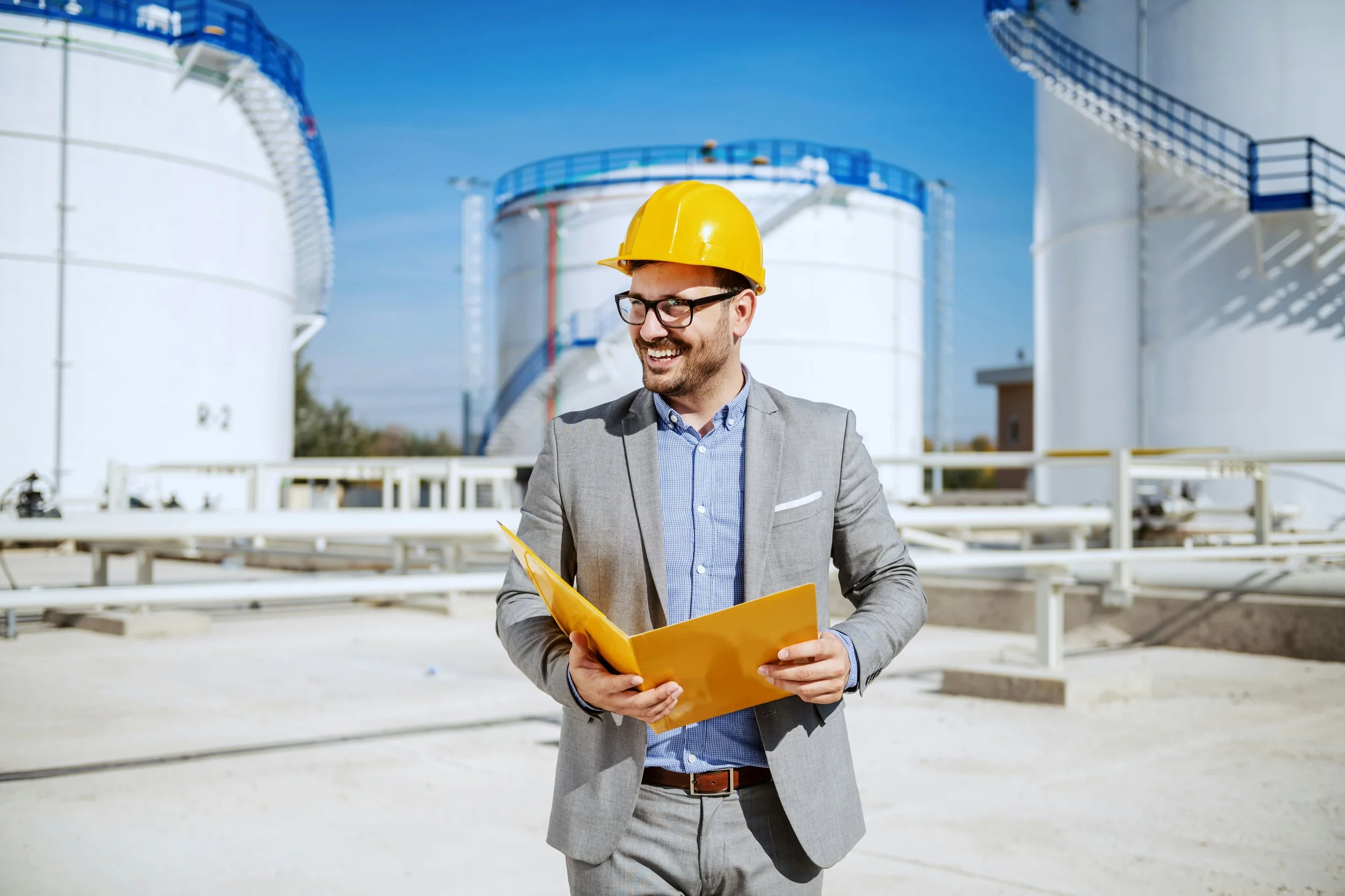 Ein Mann in einem grauen Anzug mit Krawatte und Brille, trägt einen gelben Schutzhelm und hält eine gelbe Aktenmappe, während er vor einer Industrieanlage steht, die große Tanks und Rohrleitungen umfasst.
