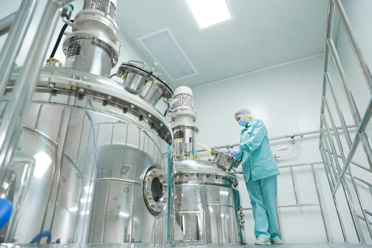 Eine Wissenschaftlerin trägt Schutzkleidung und arbeitet in einem Reinraum mit großen, glänzenden, industriellen Edelstahlgeräten.