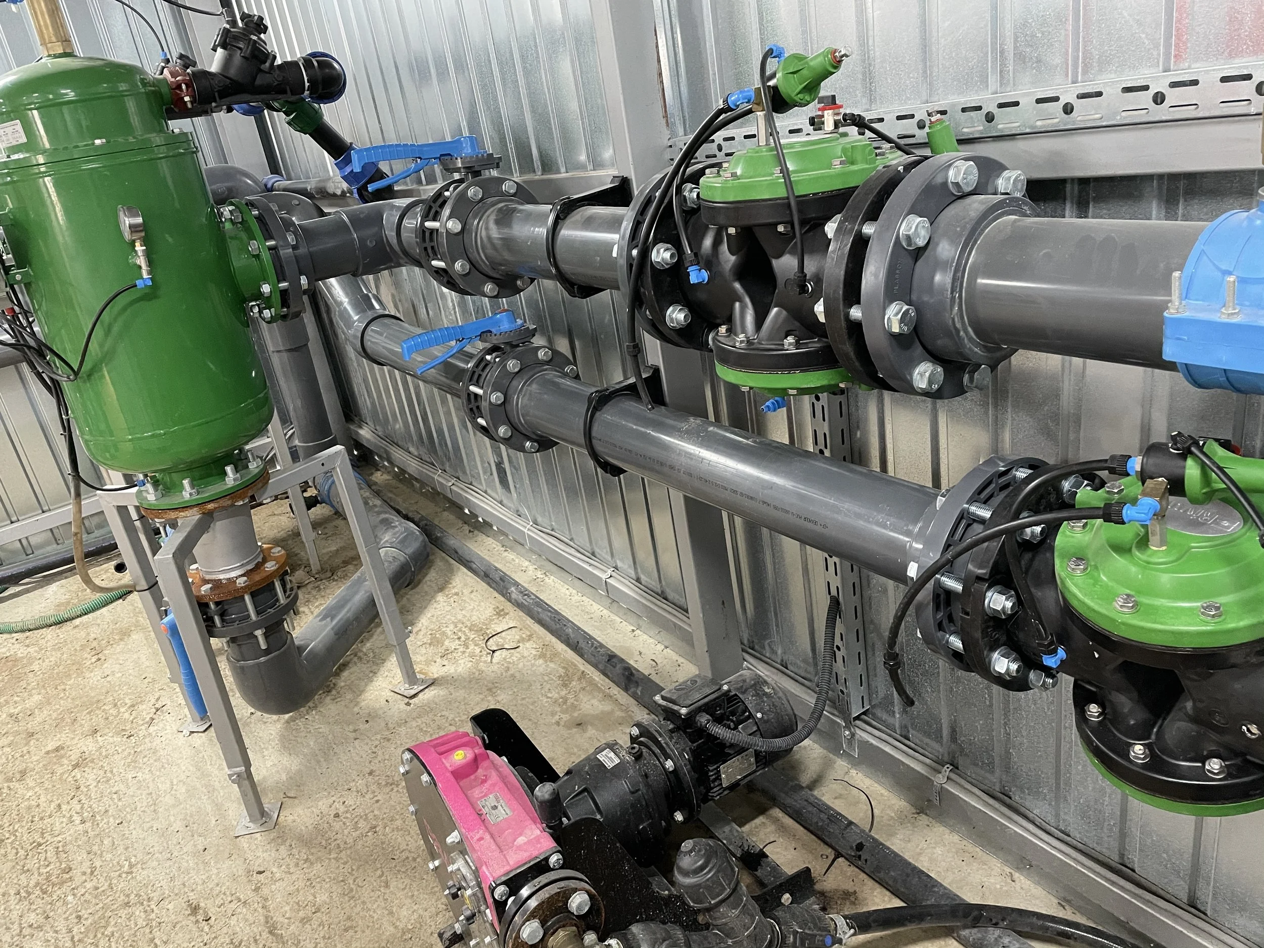 Industrial pipes and valves with green, black, and blue fittings, mounted on a metallic wall in a mechanical room.
