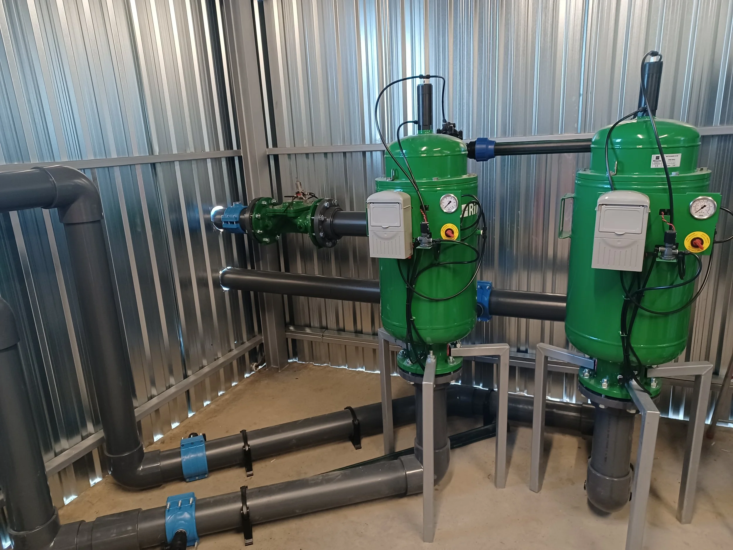 A mechanical system with green filtration units, black piping, and pressure gauges installed inside an industrial facility with metal walls.