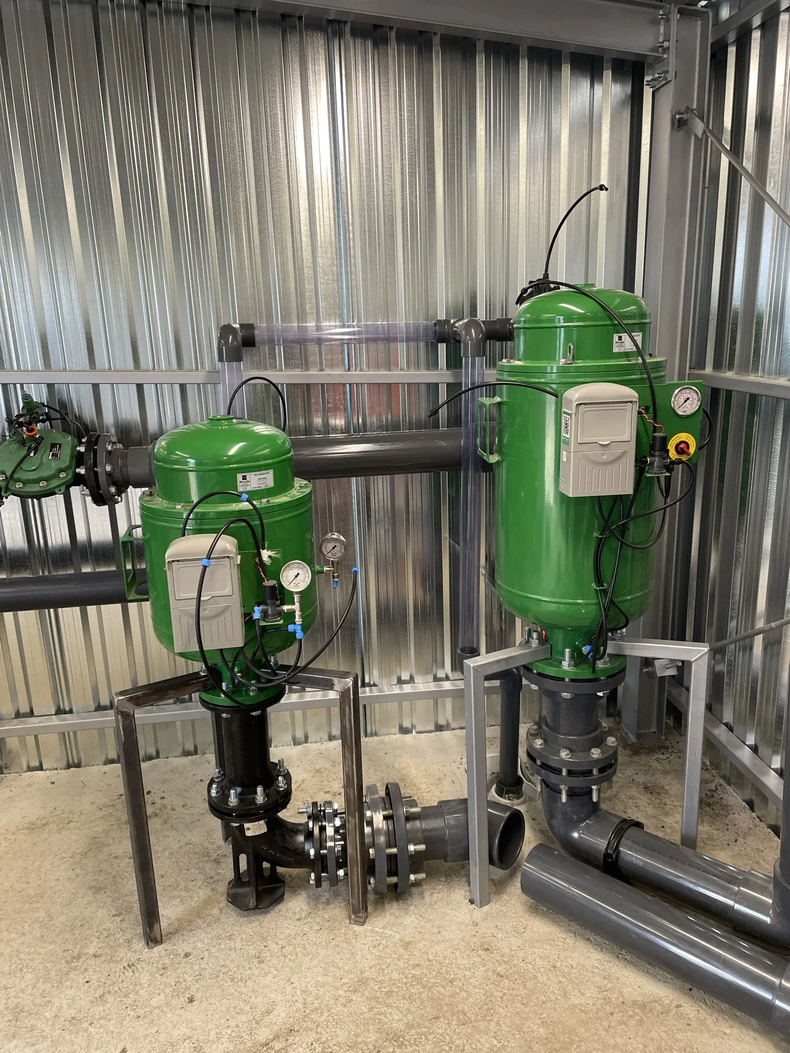 Industrial water treatment system with two green filtration units, connected pipes, gauges, and control boxes inside a metal enclosure.