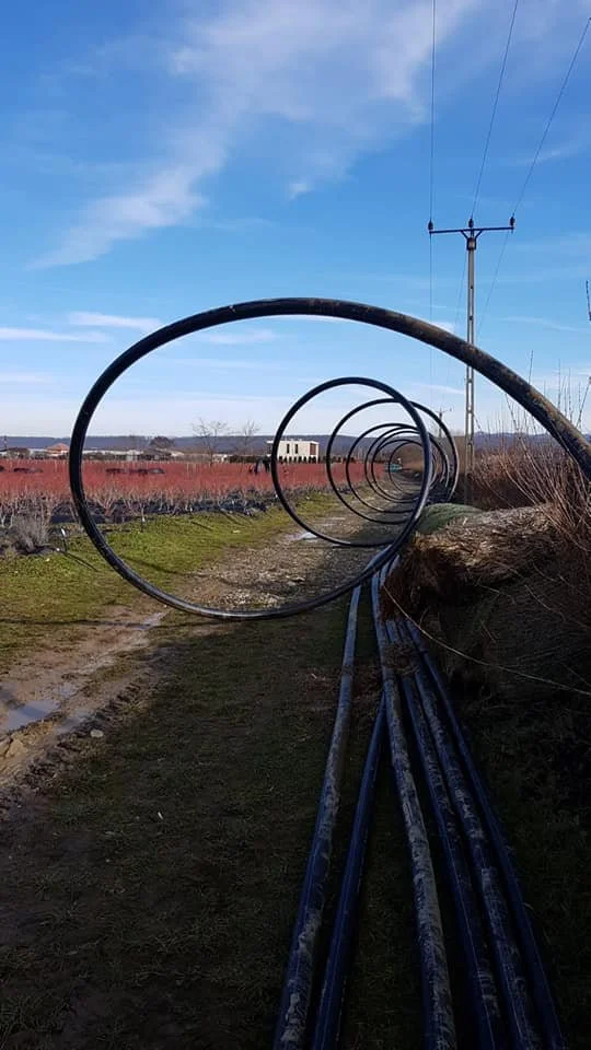 A row of large, circular metal pipes aligned along a dirt path in a rural area, with power lines and a blue sky with some clouds overhead.