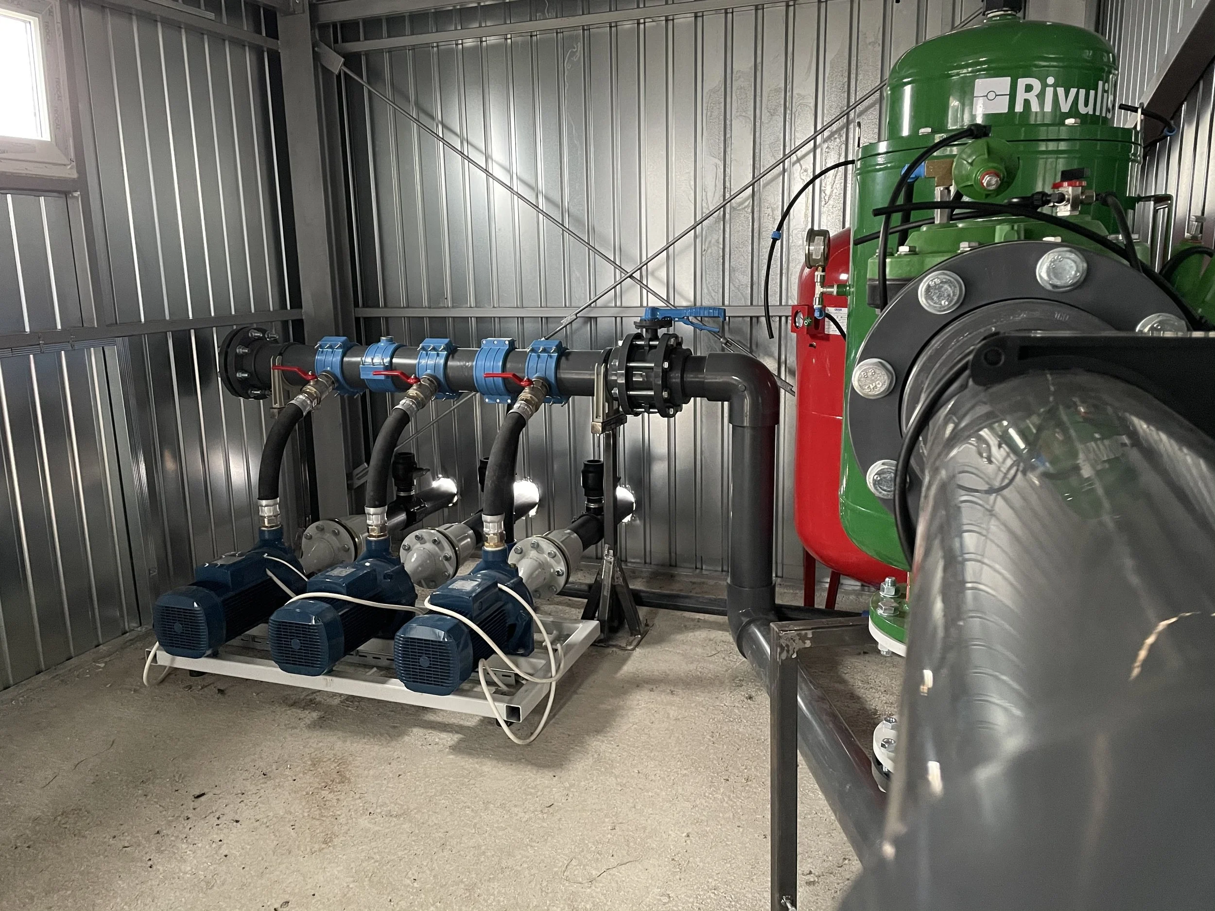 Industrial pump room with three blue pumps connected to black hoses, metal piping, and a large green and red pressure vessel in a corrugated metal room.