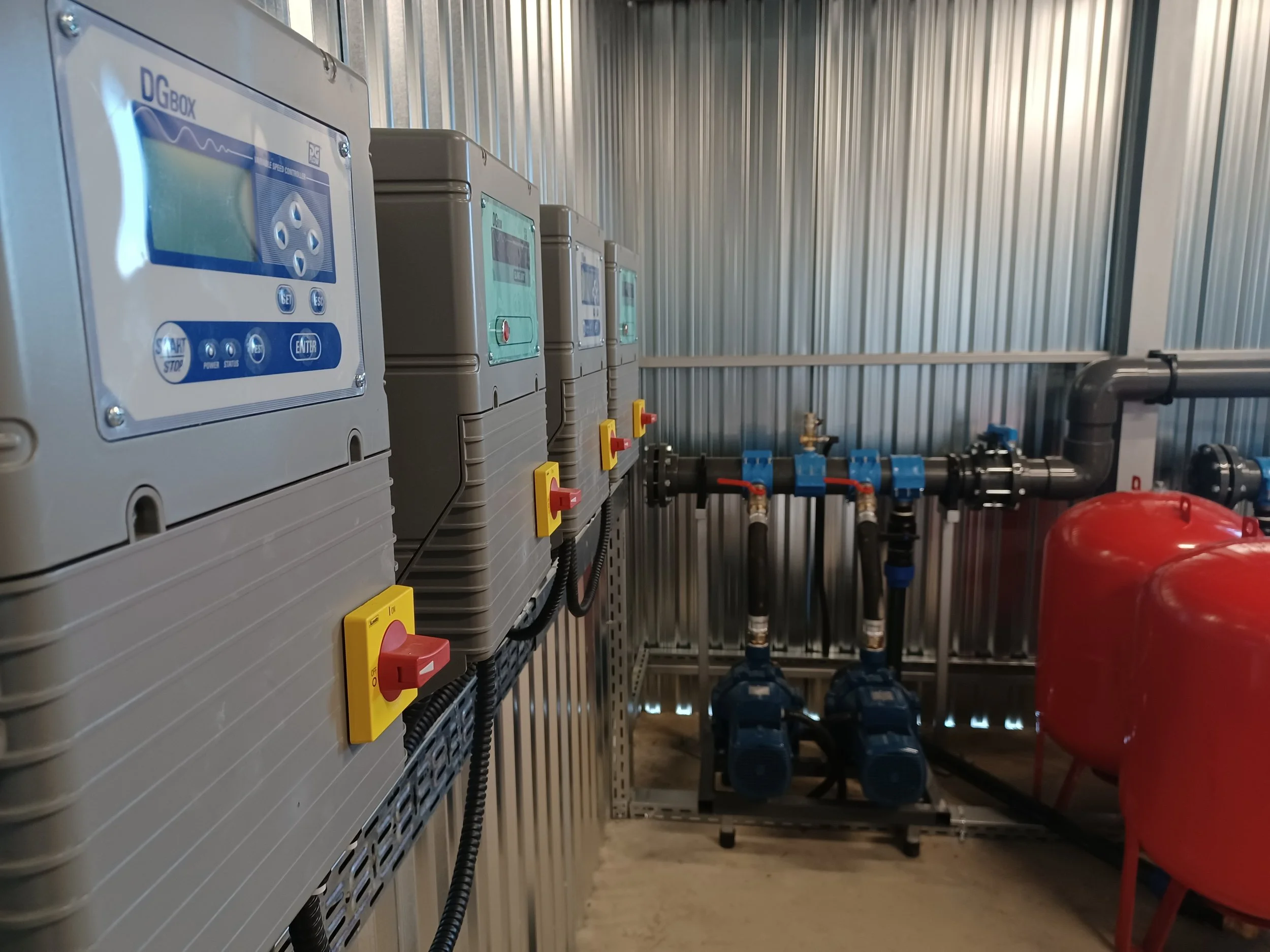 Industrial control panels mounted on a wall with yellow and red switches, connected to black and blue pipes and red tanks in a metallic room.