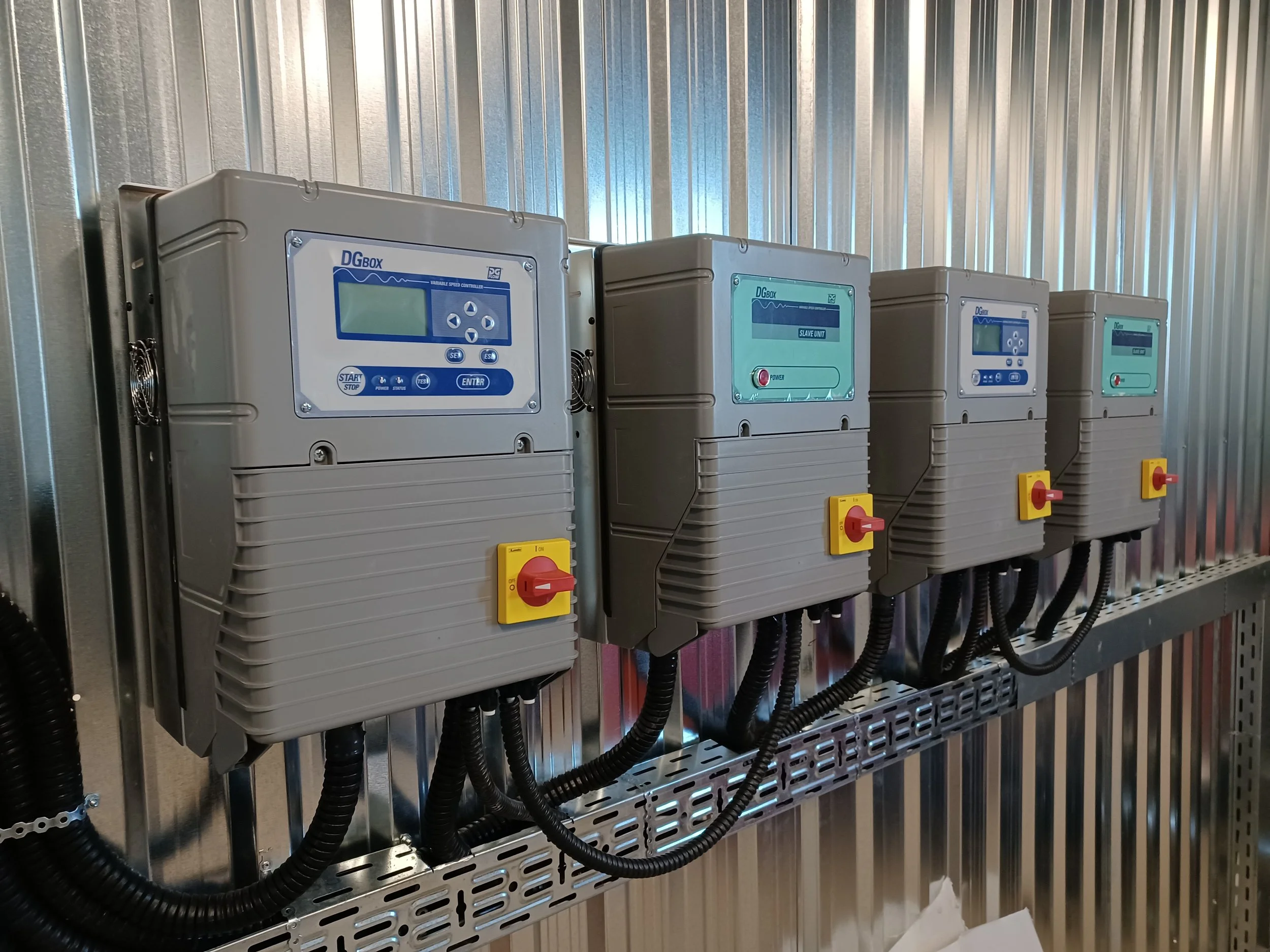 Four industrial control panels mounted on a metal wall, each with a digital display and a red and yellow switch, connected by black cables.