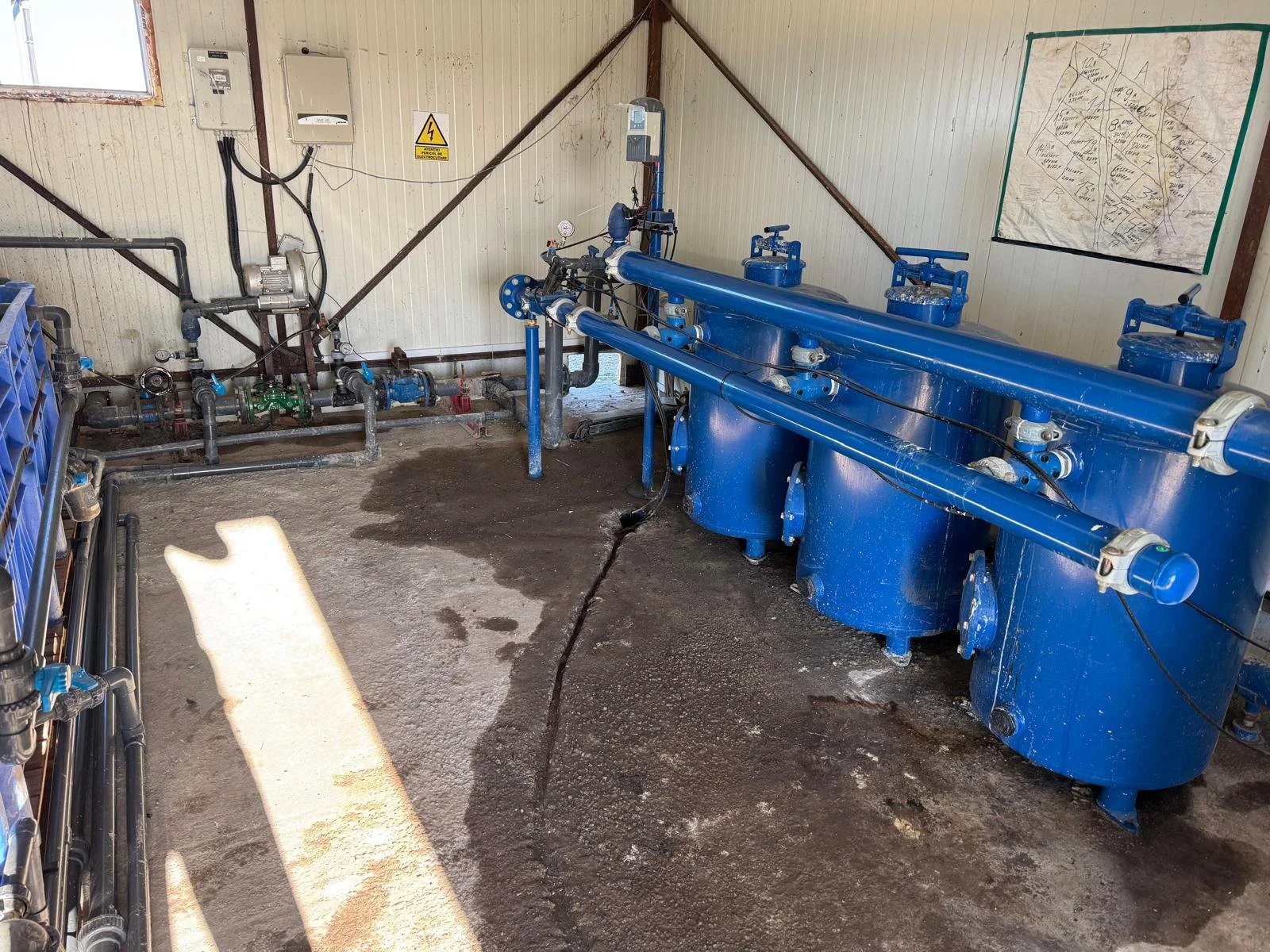 Indoor wastewater treatment equipment with blue tanks and pipes, a wall map, and electrical panels, inside a small facility.
