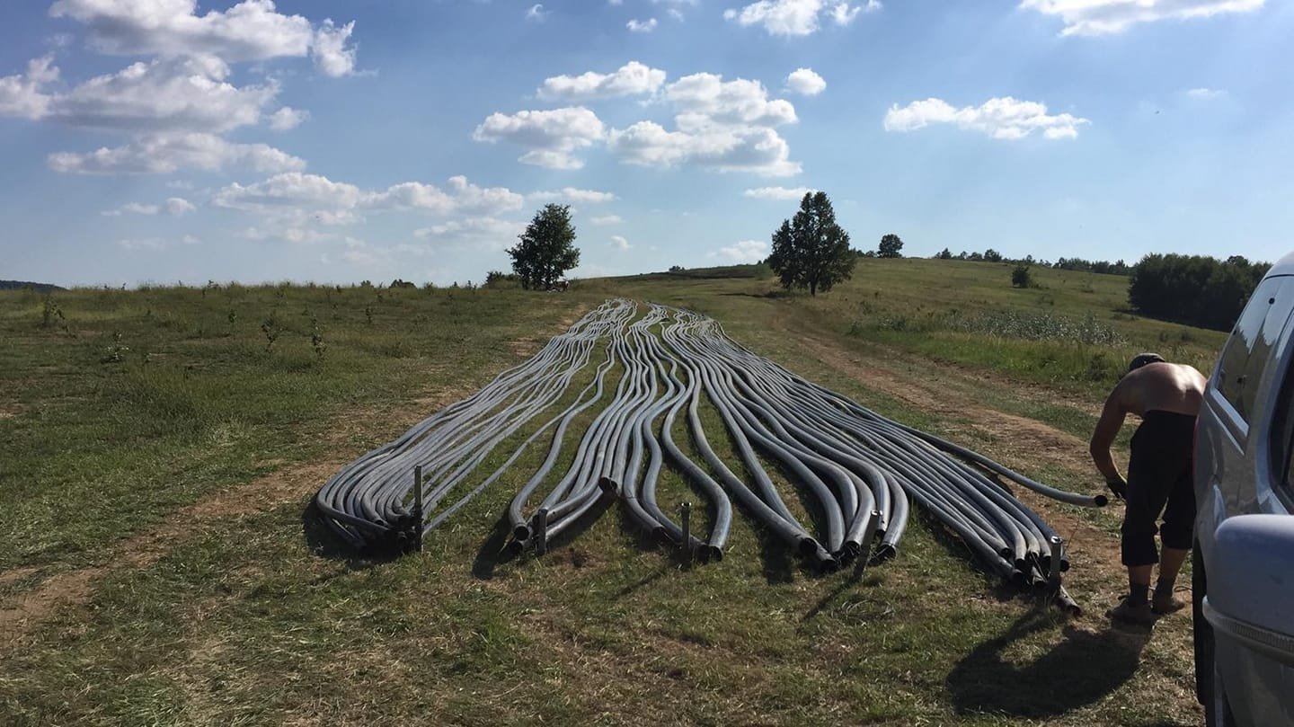 Large bundle of gray flexible pipes laid out on a grassy field, with a man bending down near a white van on the right side, in a rural landscape with trees and a blue sky with clouds.