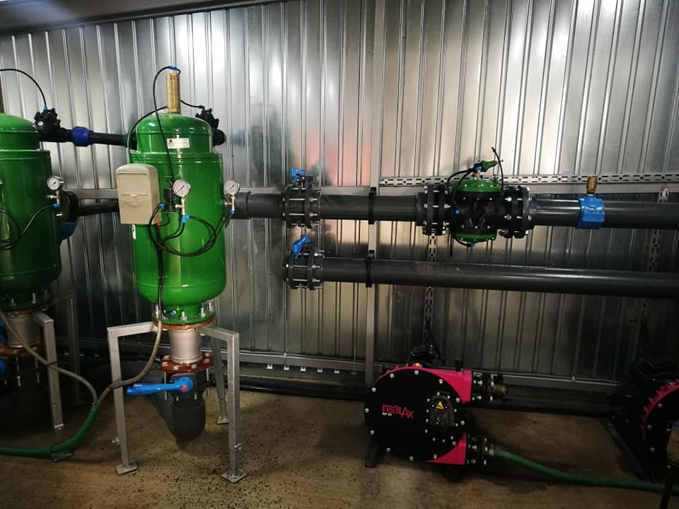 Industrial piping system with green tanks, black pipes, pressure gauges, and a red and black pump in a metallic-walled room.