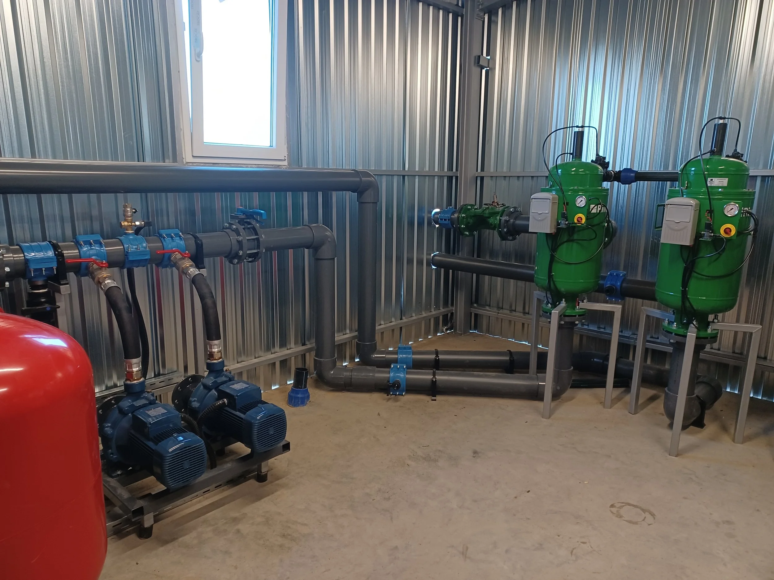 Industrial mechanical room with green filtration units, black pipes, and blue pumps against metallic walls and concrete floor.