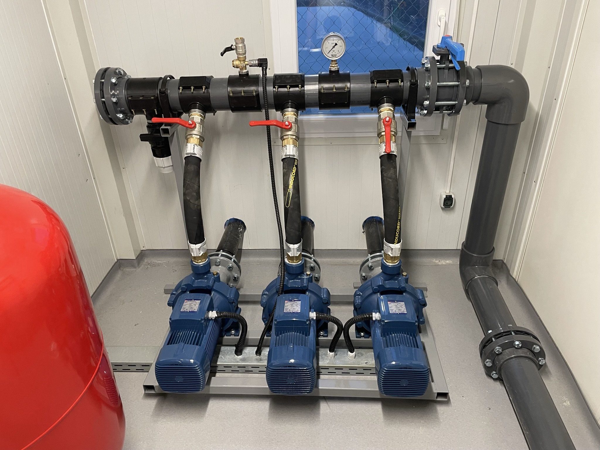 Three blue pumps connected to black pipes with red lever valves, a pressure gauge, and a black pipe on a gray floor with wall panels, a window, and an electrical socket.