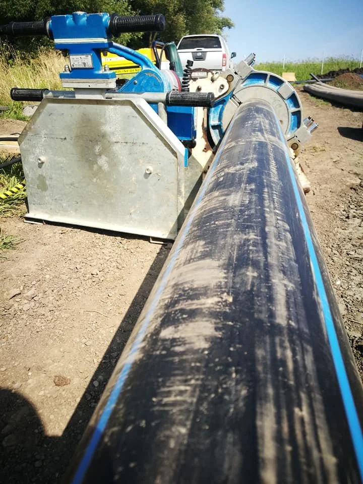 Close-up of a large industrial pipe at a construction site, with construction equipment and a white vehicle in the background.