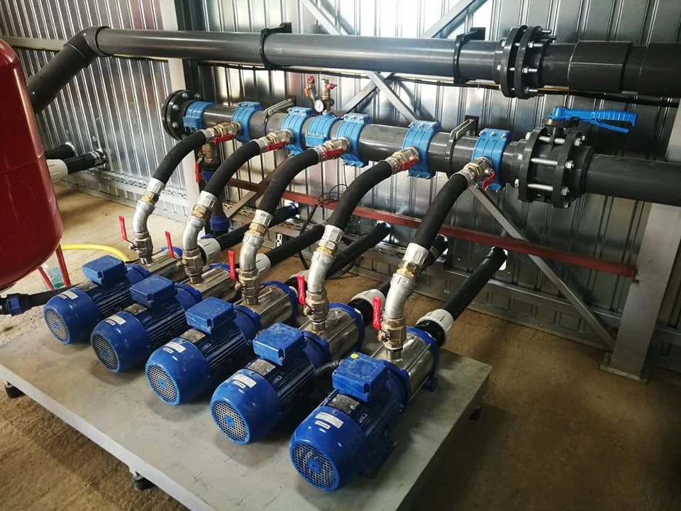 Five blue pumps connected to black pipes with red and blue valve handles, installed on a gray metal platform in an industrial setting.