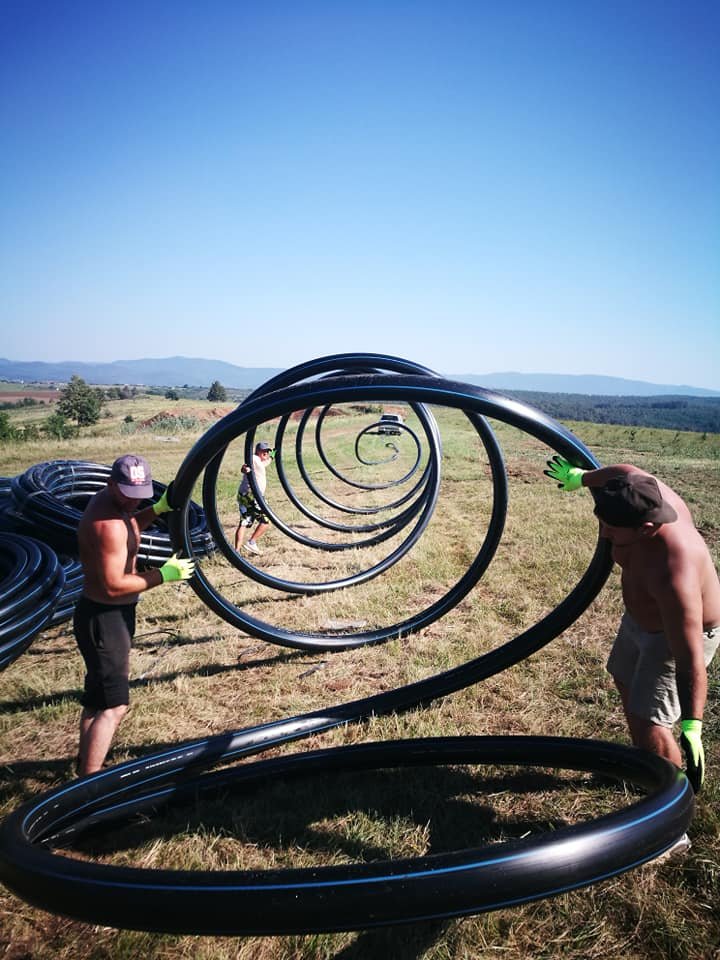 Three shirtless men wearing hats and gloves are holding large black plastic coils in an open grassy field with a clear blue sky, creating a visual illusion of a vanishing point with repetitive coils.