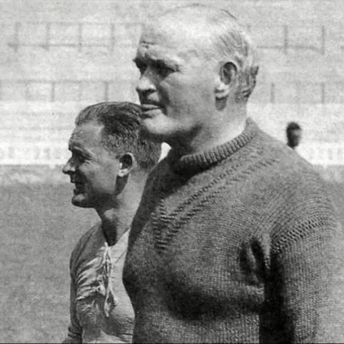 Black and white photo of two men standing outdoors, with one man in the foreground having white hair and wearing a sweater, and the other man in the background with darker hair.