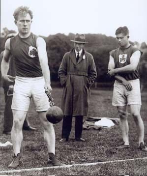 Two young male athletes in athletic uniforms standing on a field with a person in a coat and hat in the background, possibly at a sports event or track and field competition.