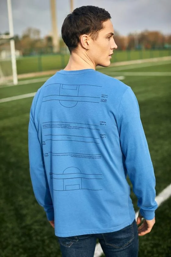 Person with short dark hair wearing a blue long-sleeve shirt with a sports field map printed on the back, standing outdoors on a grass field with goalposts in the background.