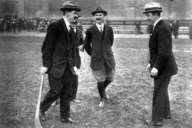 Three men in early 20th-century attire standing on a field, two of them holding hockey sticks, appearing to play or practice hockey in a park.