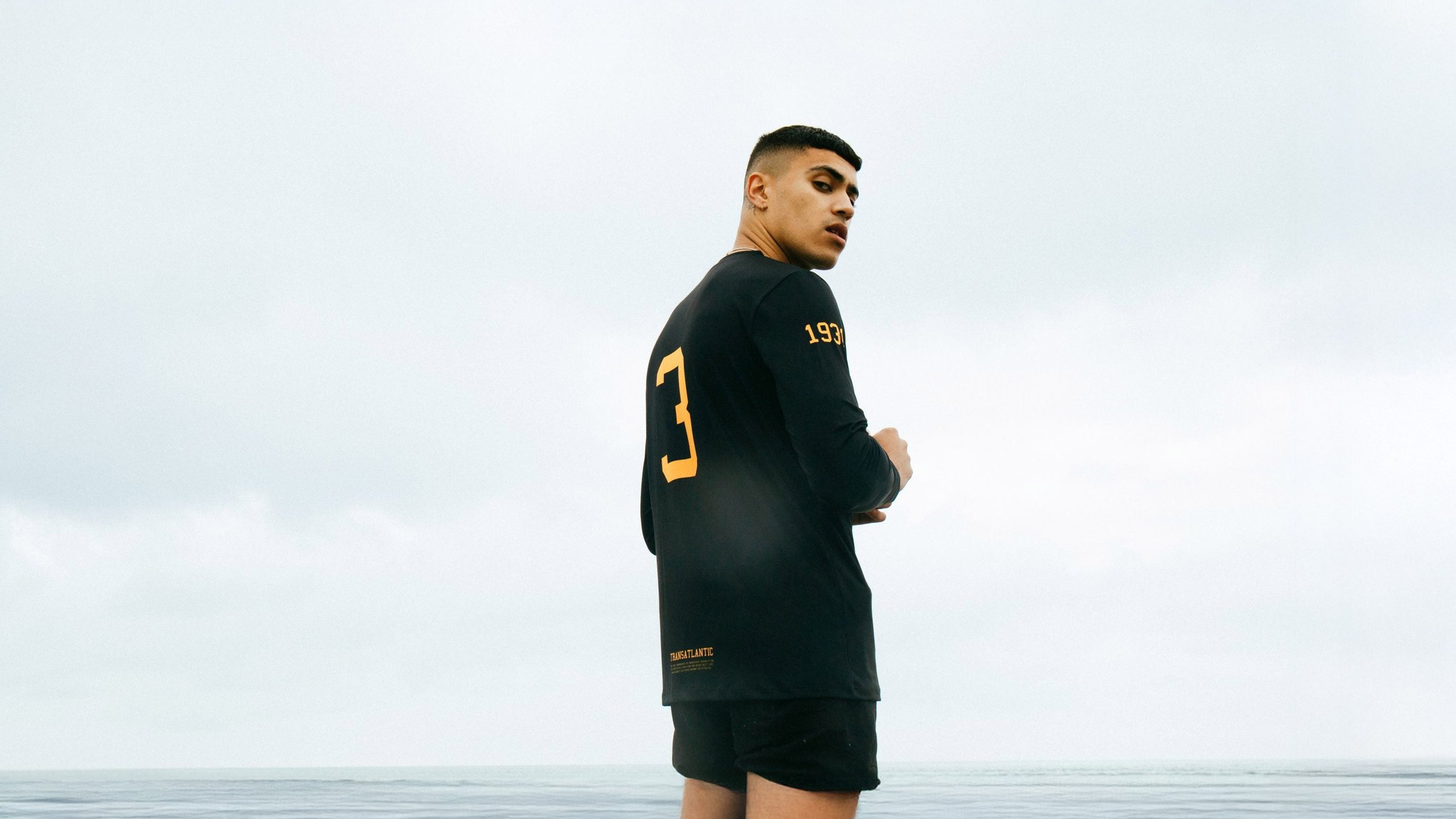 Young man standing on a beach in a black shirt with yellow lettering, looking over his shoulder at the camera, with ocean and cloudy sky in the background.