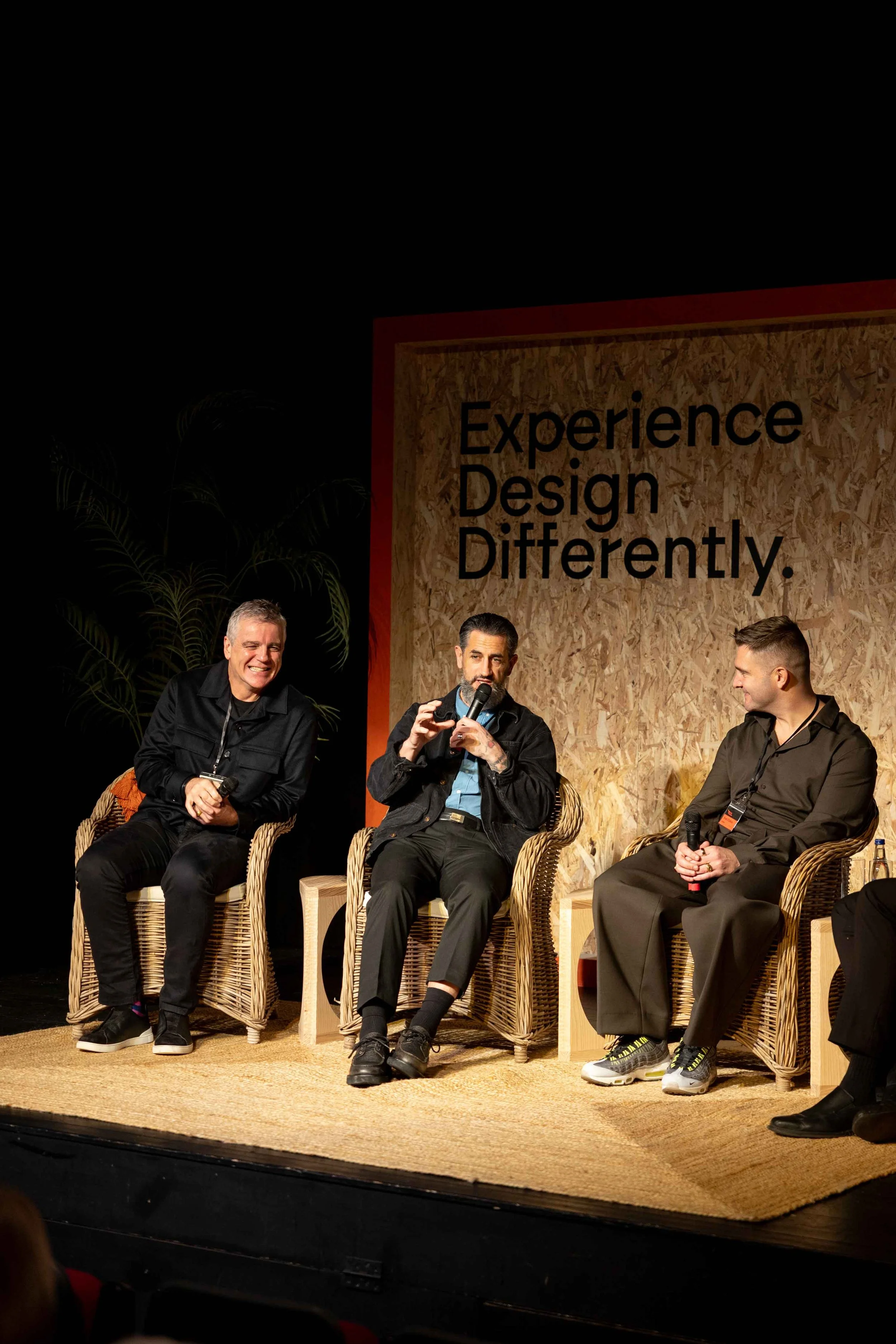 Three men sitting on stage chairs during a panel discussion at Design Kenmare festival with a sign behind them that says, "Experience Design Differently."
