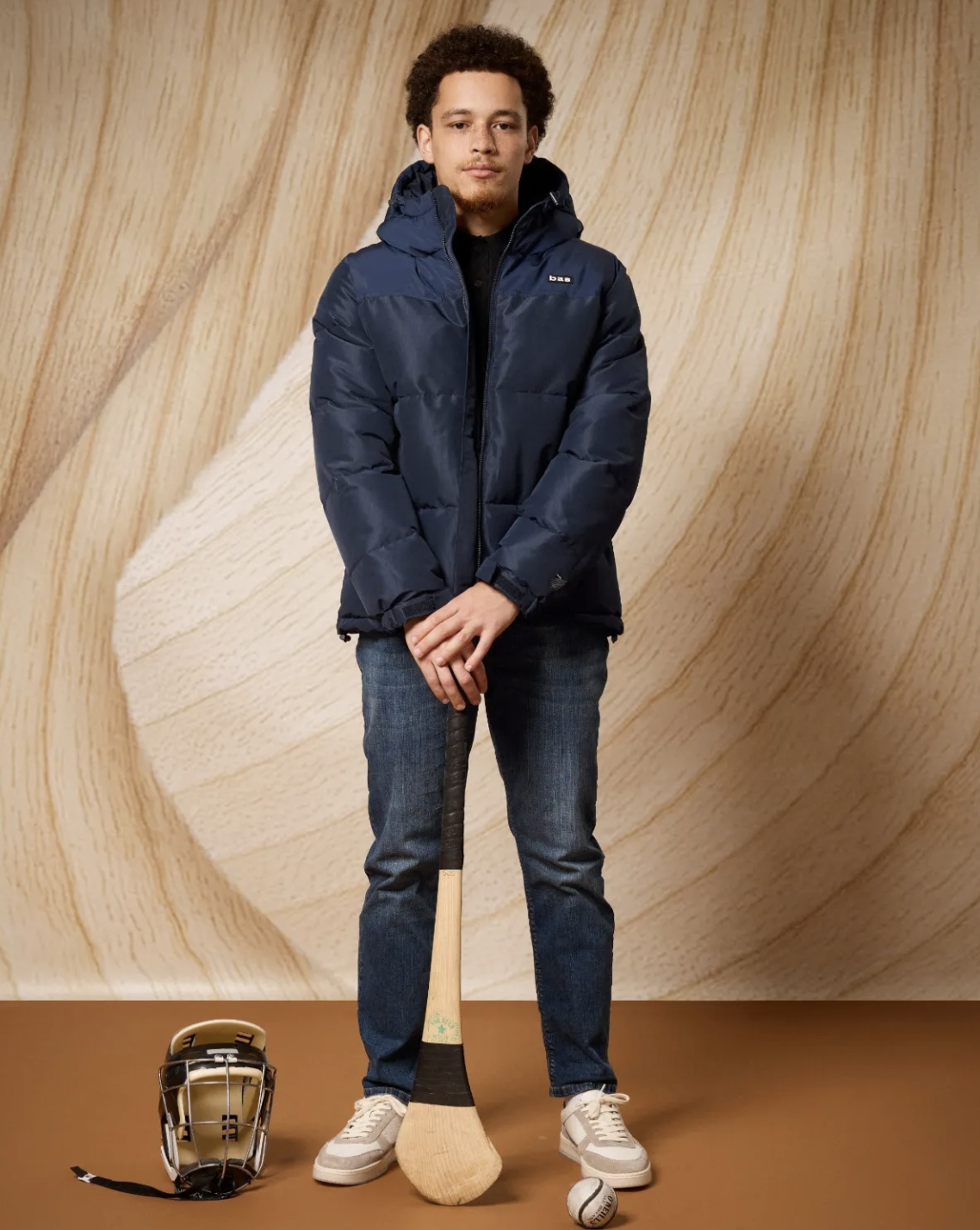 A young man stands in front of a wooden background with a hockey helmet, a hockey stick, and a baseball on the floor, dressed in casual clothing with a blue puffer jacket.