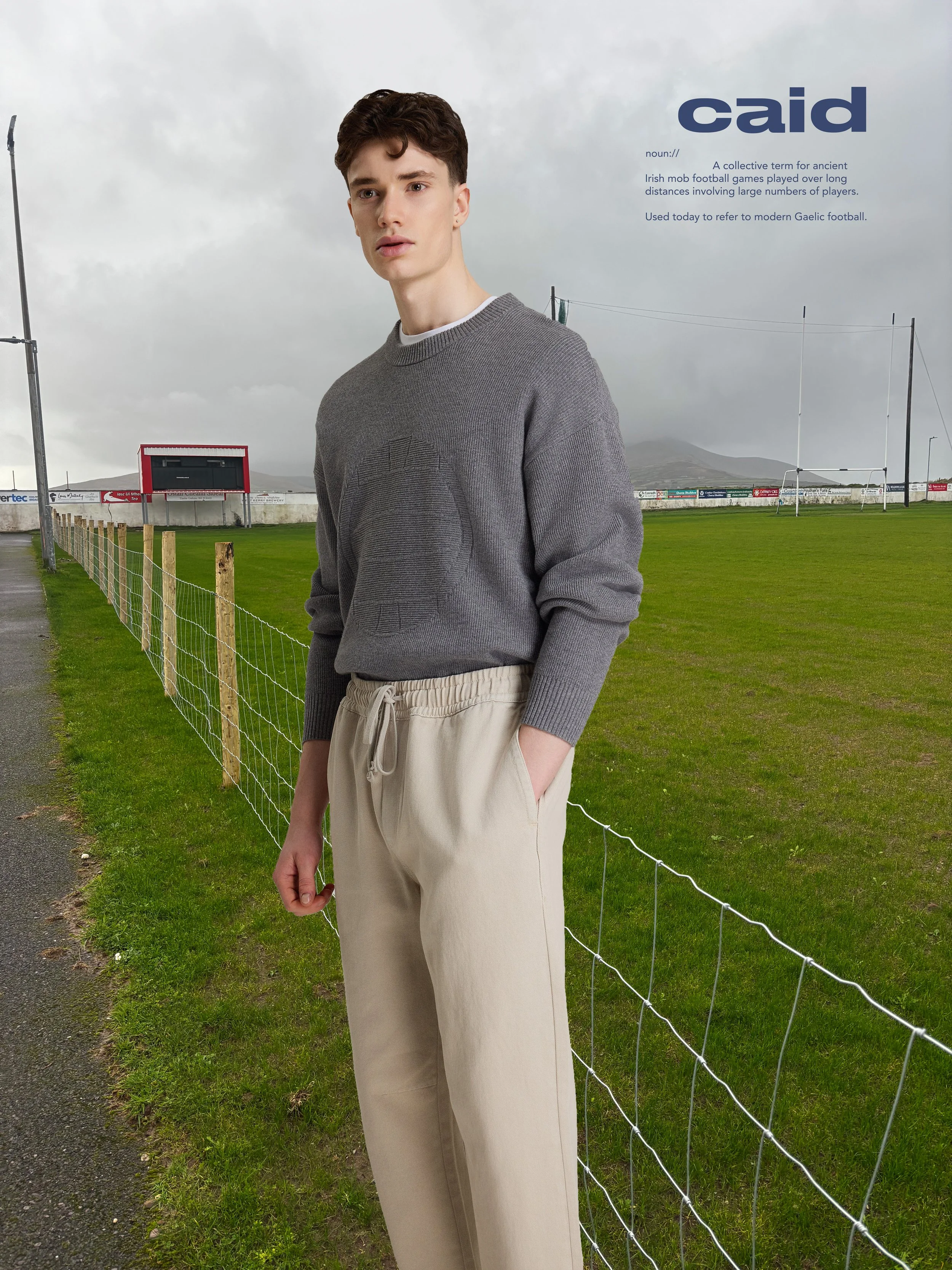 A young man with wavy dark hair, wearing a gray sweatshirt and beige pants, standing outdoors near a fenced grassy field with rugby goalposts and a scoreboard in the background under a cloudy sky.
