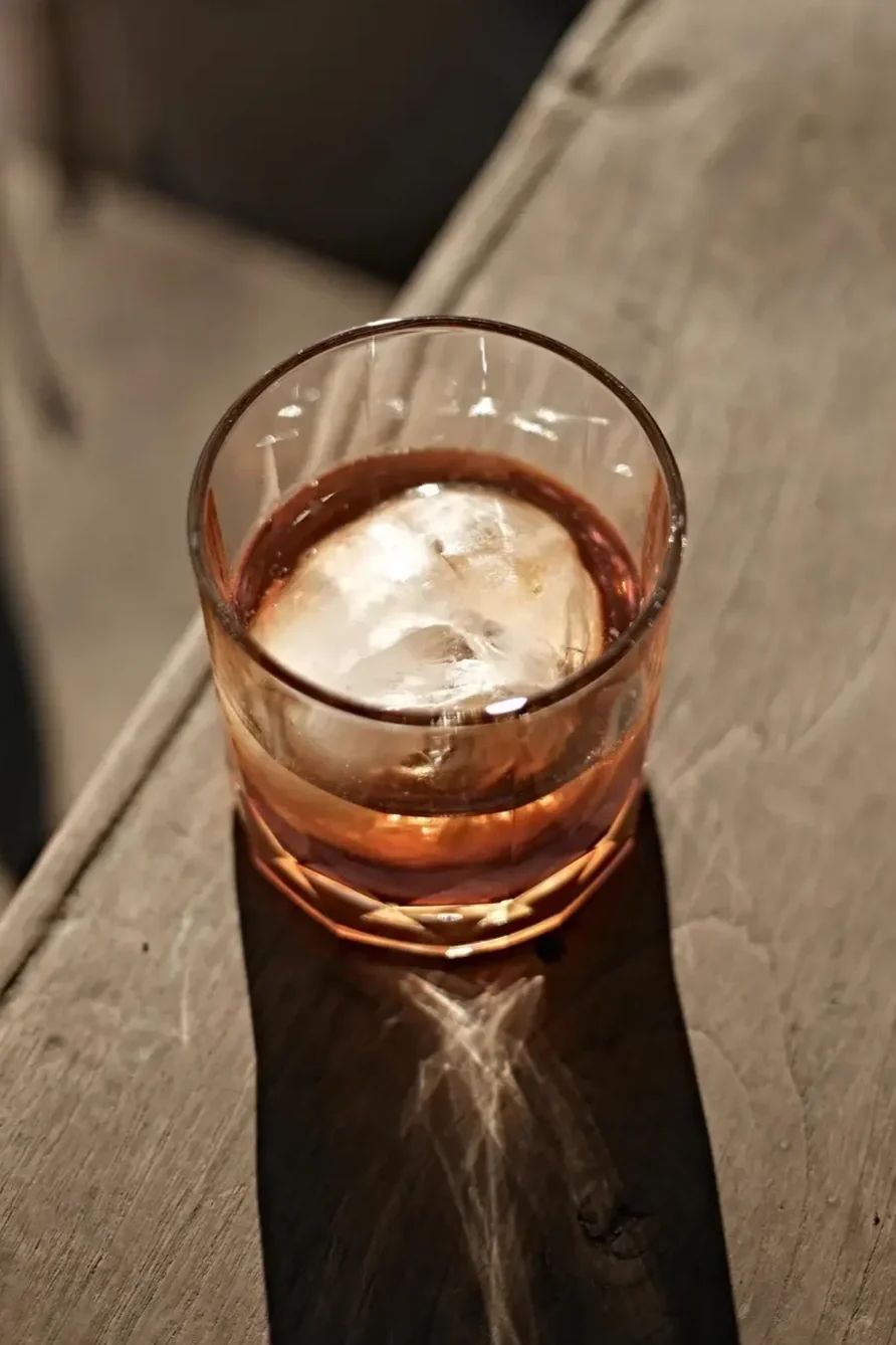 Verre contenant une boisson sombre avec un gros glaçon, posé sur une surface en bois, avec un éclairage créant une ombre et un reflet sur la table.