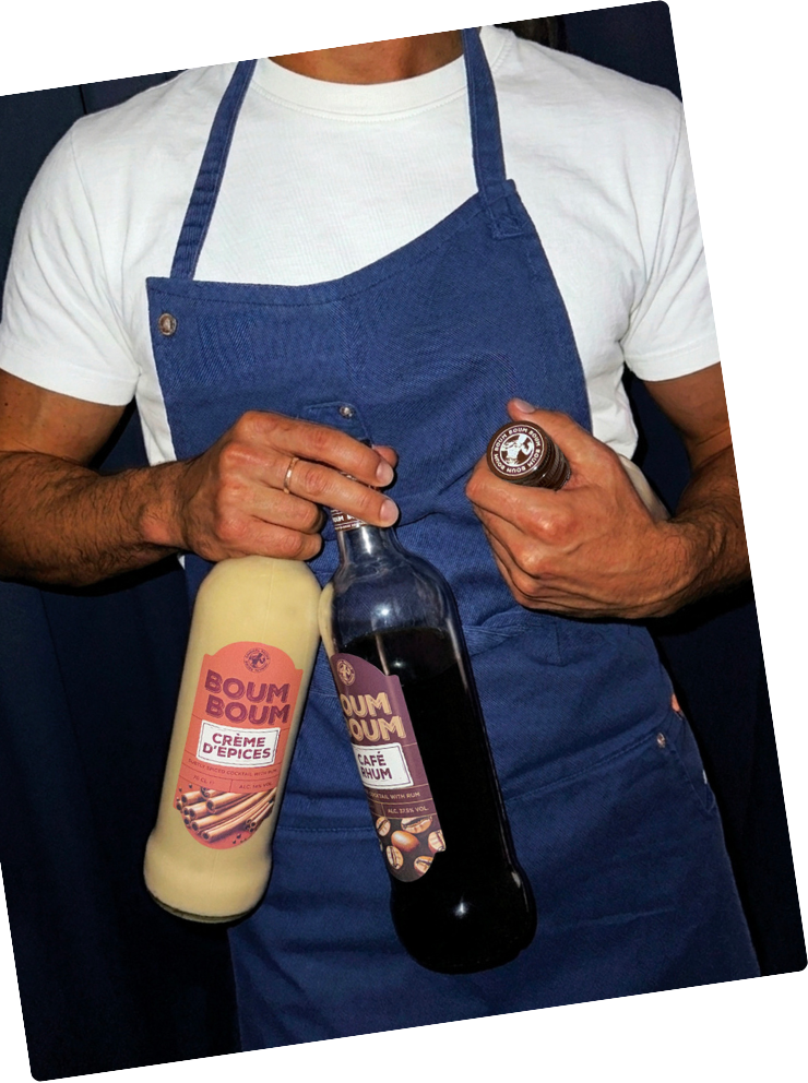 Un homme portant un tablier bleu, tenant deux bouteilles, une de crème brûlée épicée et une de café au rhum.