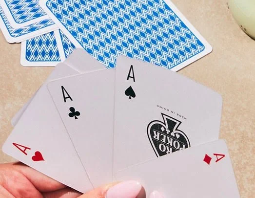 Quatre cartes à jouer : un as de cœur, un as de trèfle, et deux autres cartes face visible. Des cartes à jouer retournées avec un dos bleu et blanc à motifs zigzag sont visibles en arrière-plan.