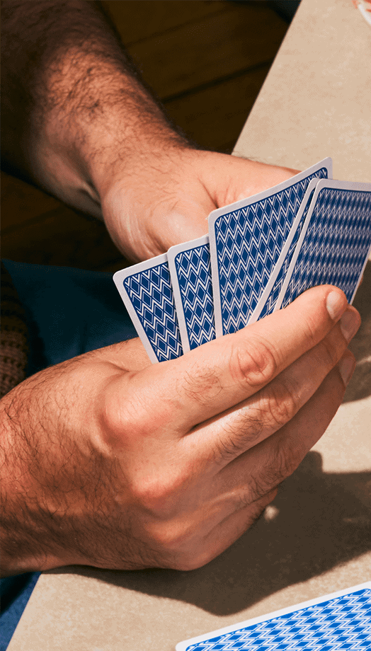 Main tenant des cartes à jouer face cachée, montrant le dos bleu et blanc, alors qu'une autre personne observe ou joue à un jeu de cartes.