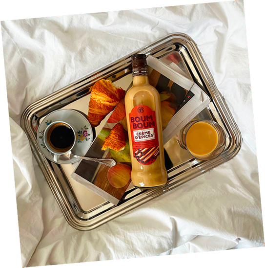 Plateau avec une bouteille de crème d'épices, un verre de jus d'orange, un petit bol de café avec une cuillère, et des croissants, posé sur une surface en tissu blanc.