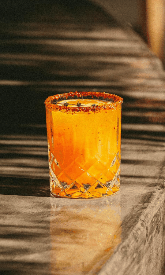 Un verre de cocktail orange avec un rebord épicé, placé sur une surface en bois. En arrière-plan, un fond flou avec des textures horizontales.