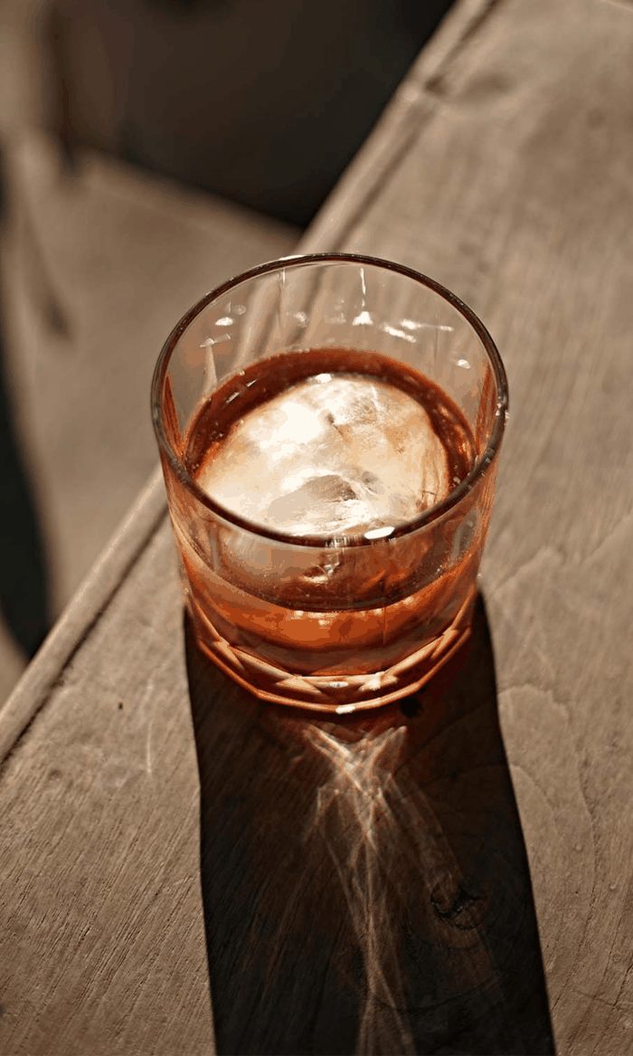 Verre de boisson avec glaçons sur une table en bois, lumière créant un reflet au fond du verre.
