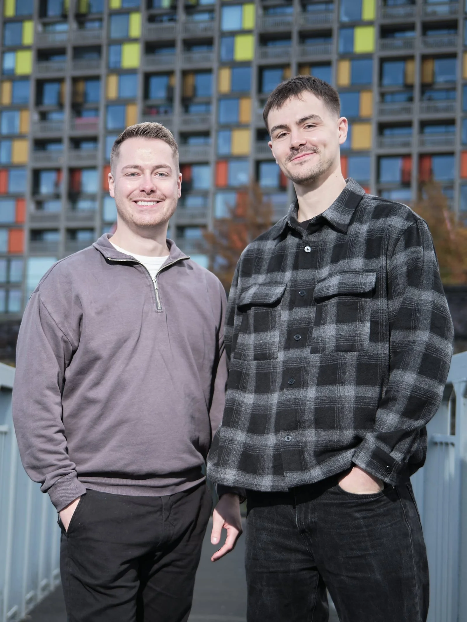 Two young men standing outdoors in front of a colorful building with squares of blue, yellow, and red, smiling at the camera.