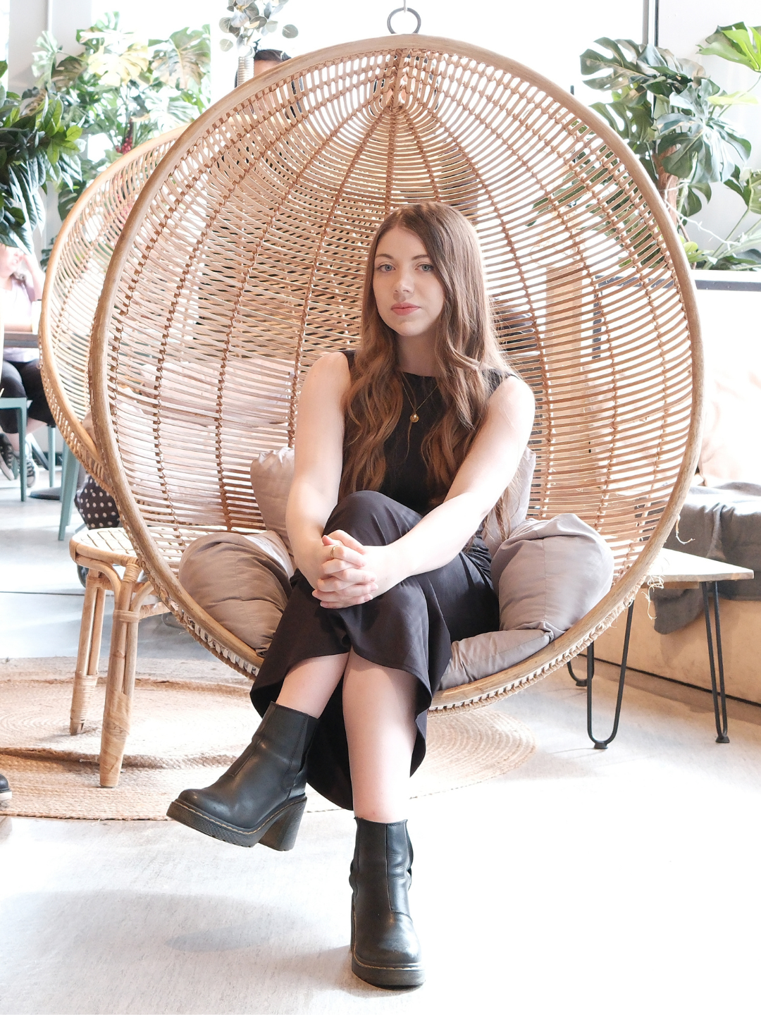 Young woman with long brown hair sitting in a round woven rattan hanging chair in a bright indoor space with green plants and other seating areas.