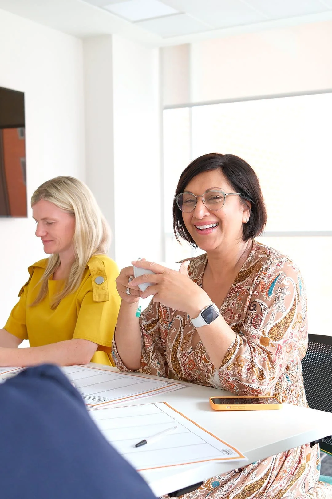 Two women sitting at a table during a meeting, smiling and talking, with documents and a smartphone on the table.