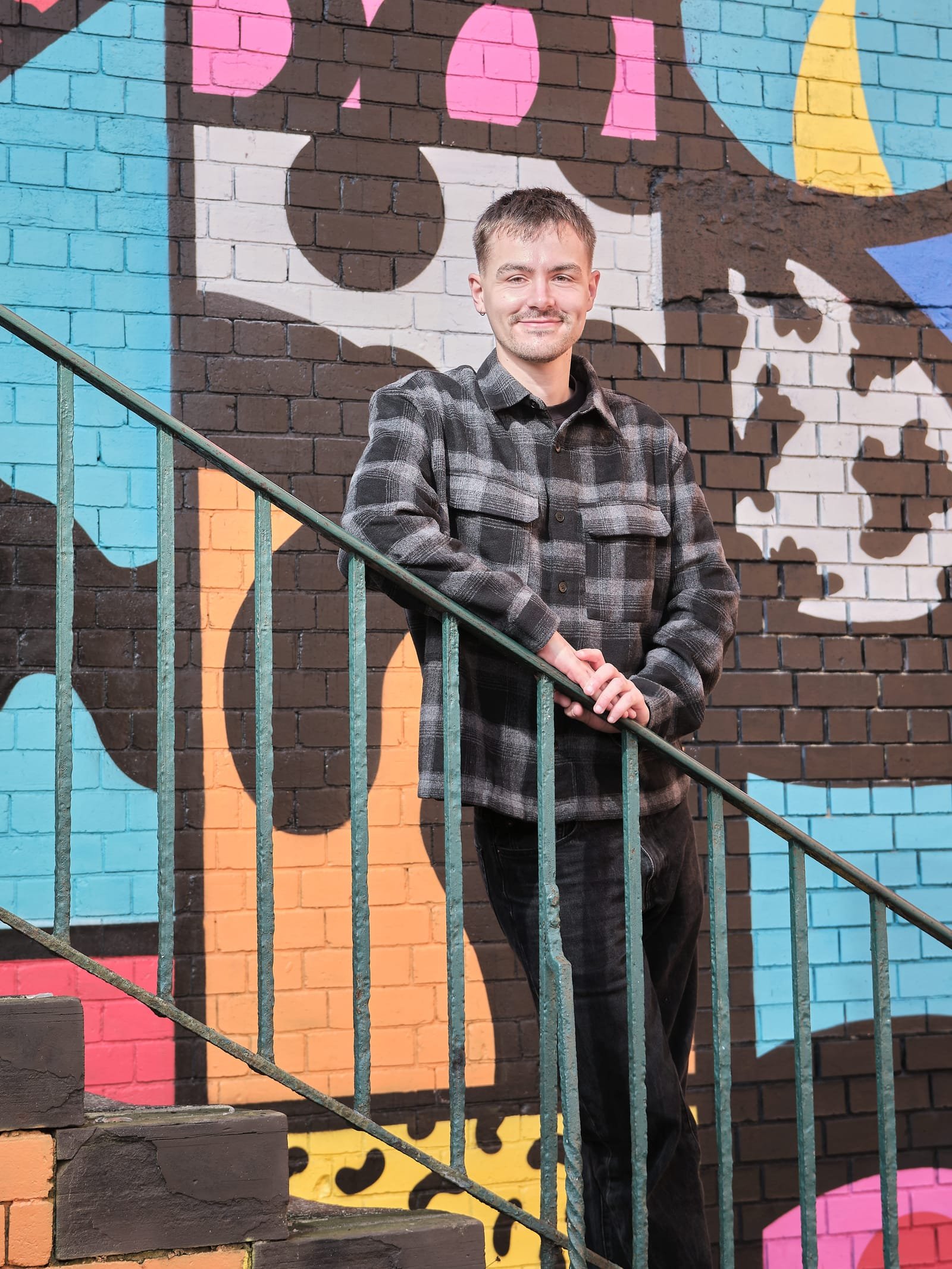 A young man stands on staircase with a colorful mural behind him, smiling at the camera.