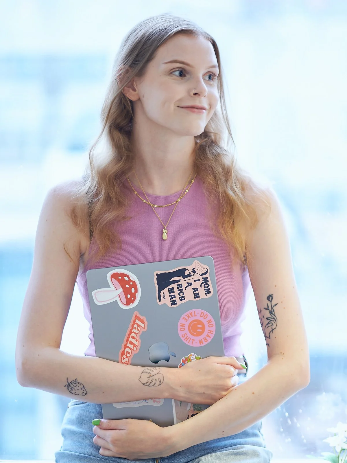 A young woman with light brown hair and tattoos on her arms, holding a laptop decorated with stickers, standing in front of a bright window.