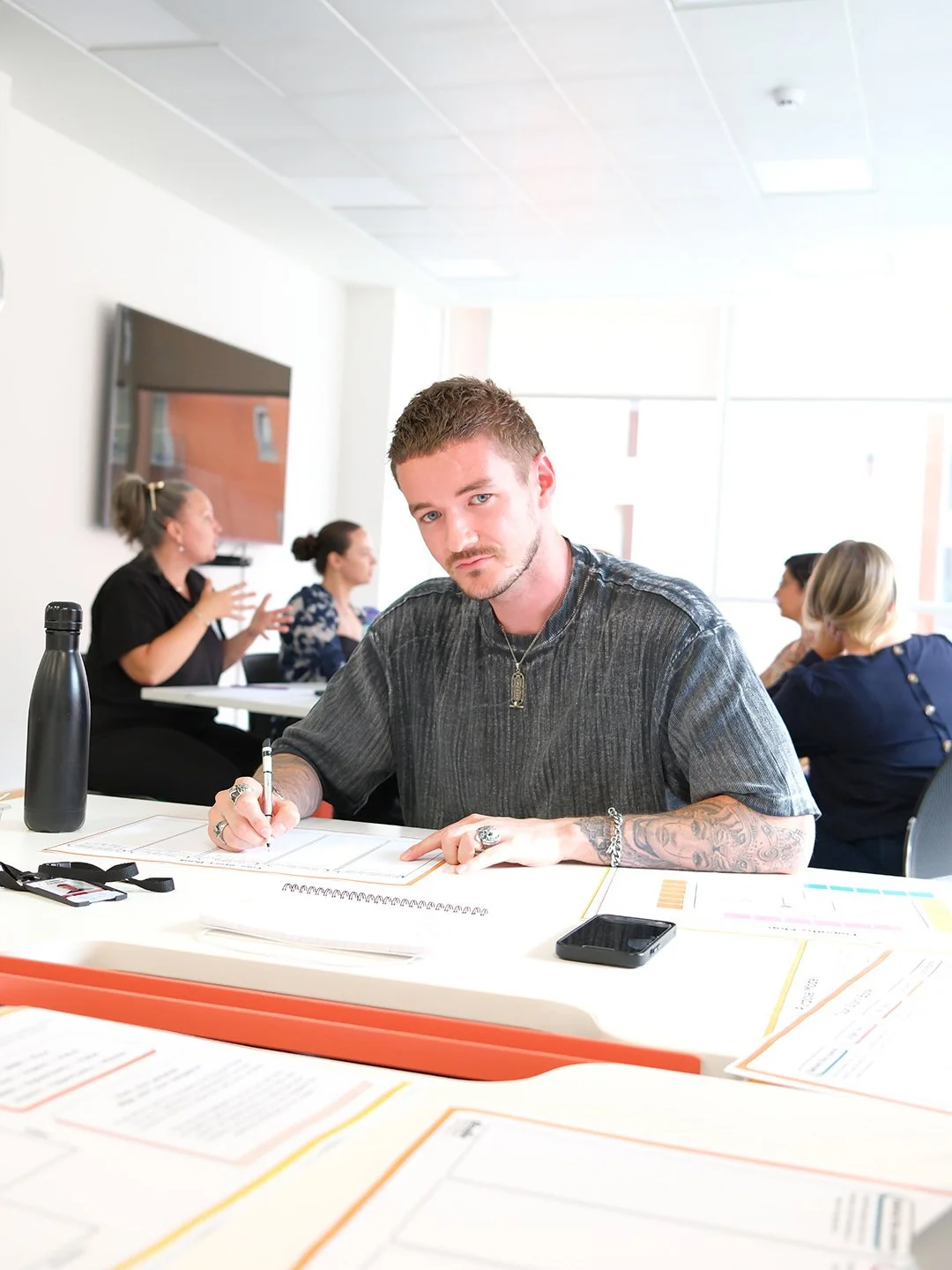 A young man with tattoos, wearing a black shirt, sitting at a table with papers, a smartphone, and a water bottle, in a bright conference room, with women in the background engaged in conversation.