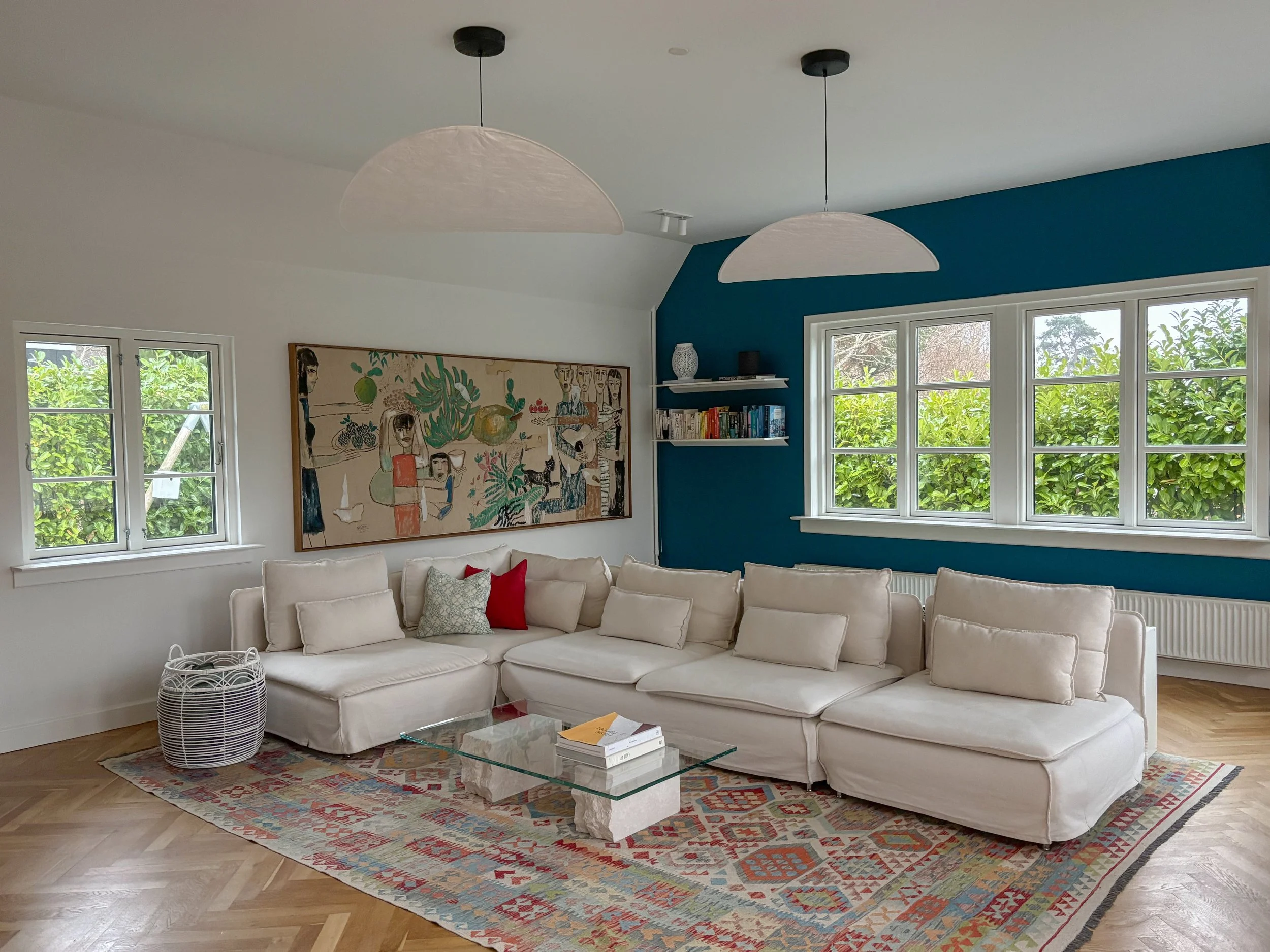 Bright living room with large white sectional sofa, colorful area rug, glass coffee table, artwork on the wall, and multiple windows with greenery outside.