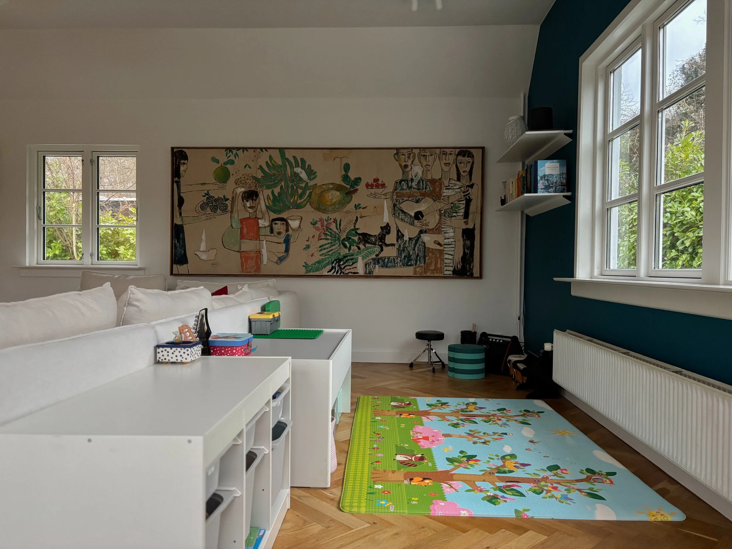 Living room with white couch, colorful children's playmat, large artwork on the wall, windows showing outdoor greenery, and various shelves and objects.