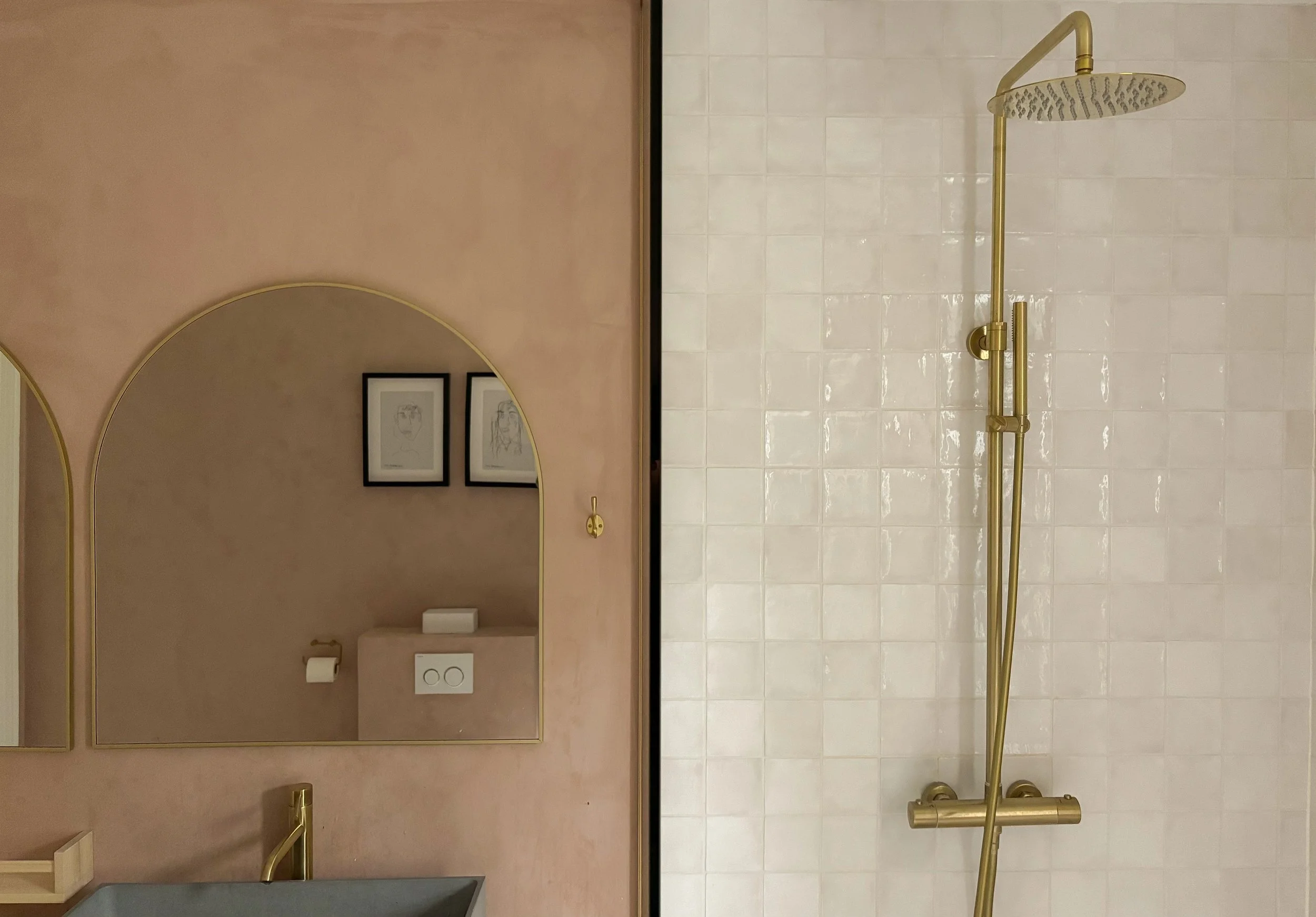 Split view of a bathroom with a pink wall and gold accents. On the left, a bathroom mirror reflects framed line art sketches on the wall and a toilet paper holder. On the right, a gold showerhead with a handheld attachment is mounted on white tiled wall.