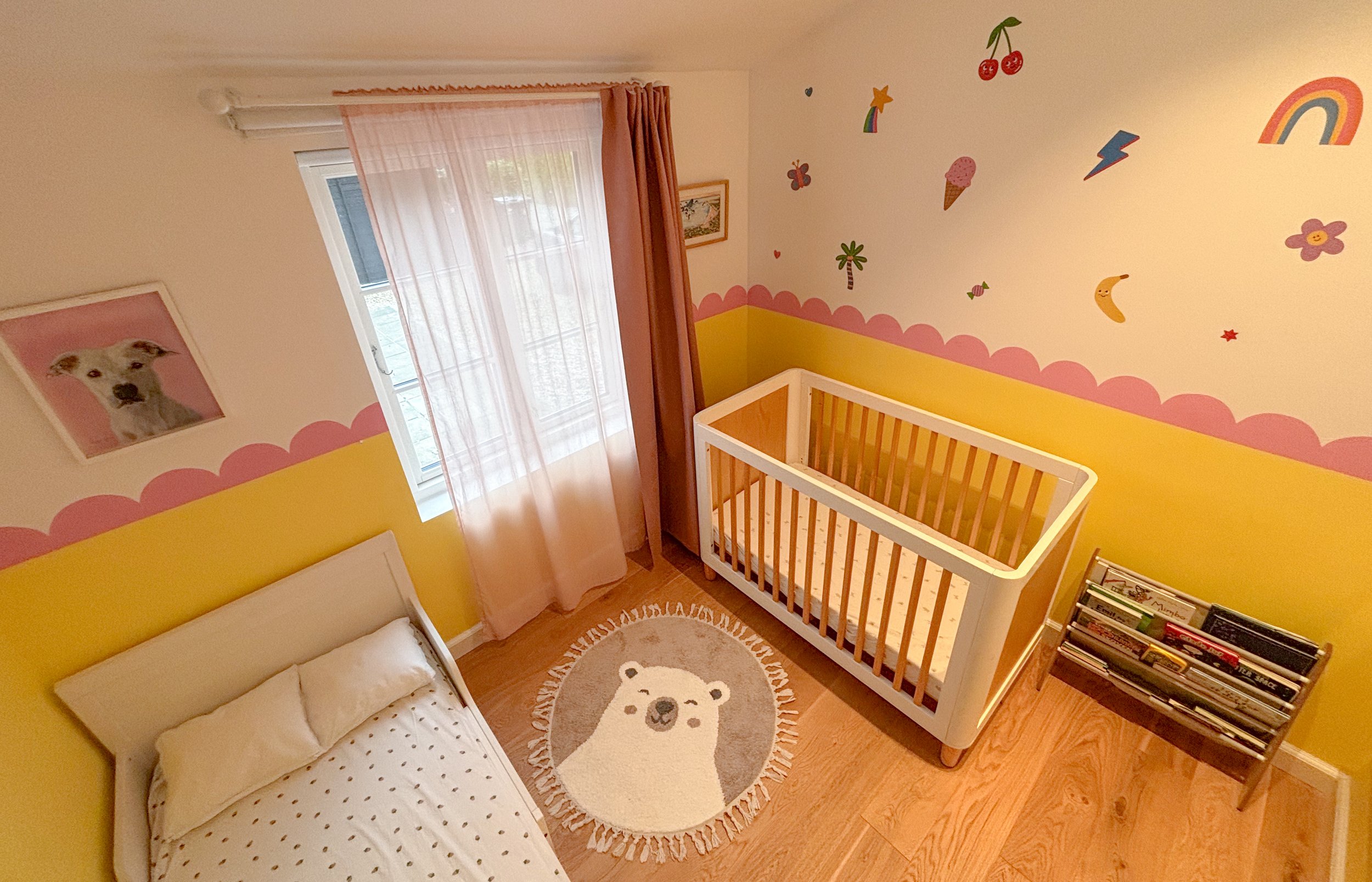 A colorful child's bedroom with pink, yellow, and white walls decorated with stickers of a rainbow, cherries, ice cream, clouds, stars, butterflies, flowers, and a banana. The room has a white bed with polka dot sheets, a white crib, a round rug with
