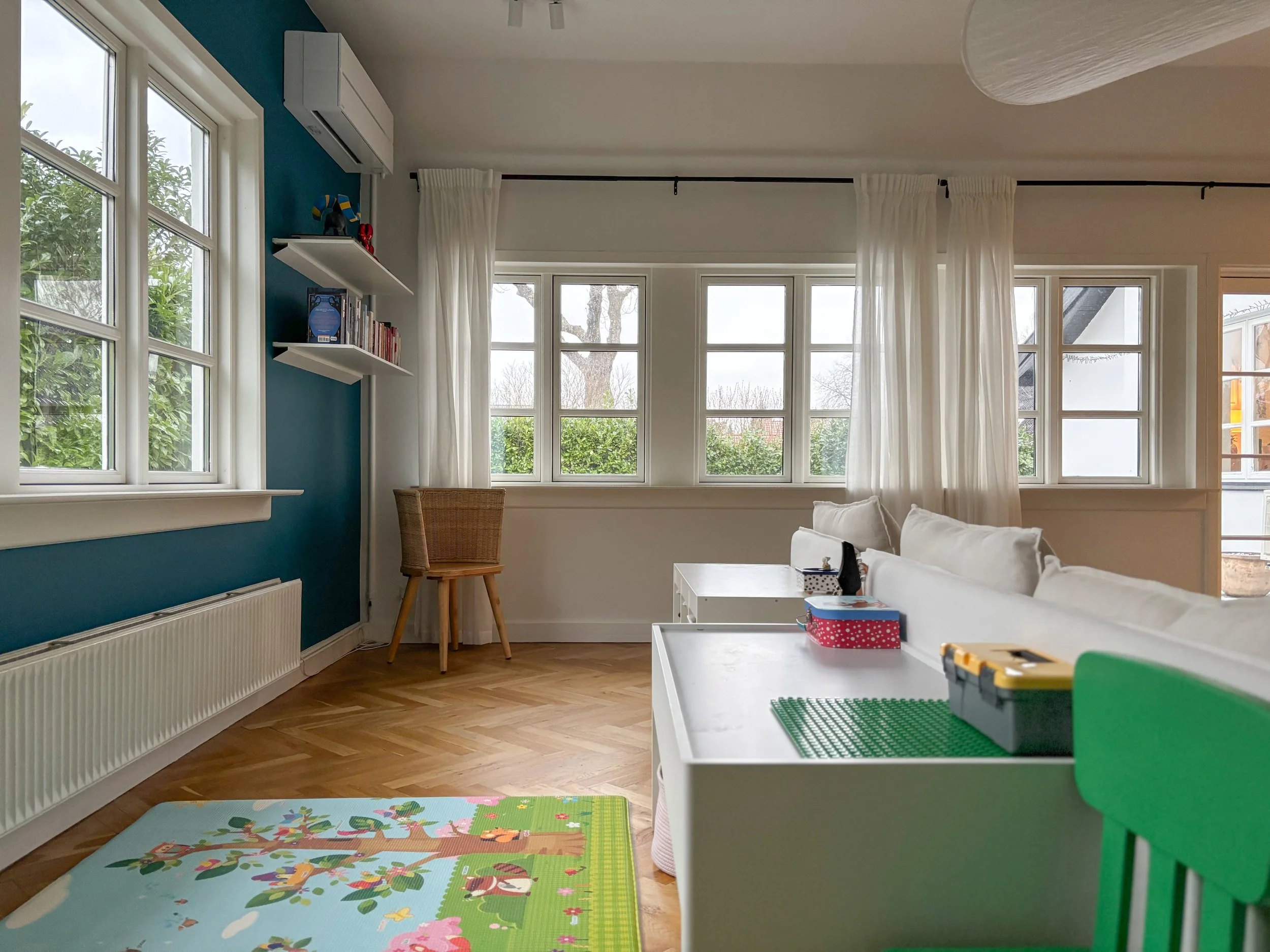 Bright living room with large windows, white curtains, a white sofa, a bamboo chair, a colorful playmat for children, a white shelf with toys and books, and a green chair.